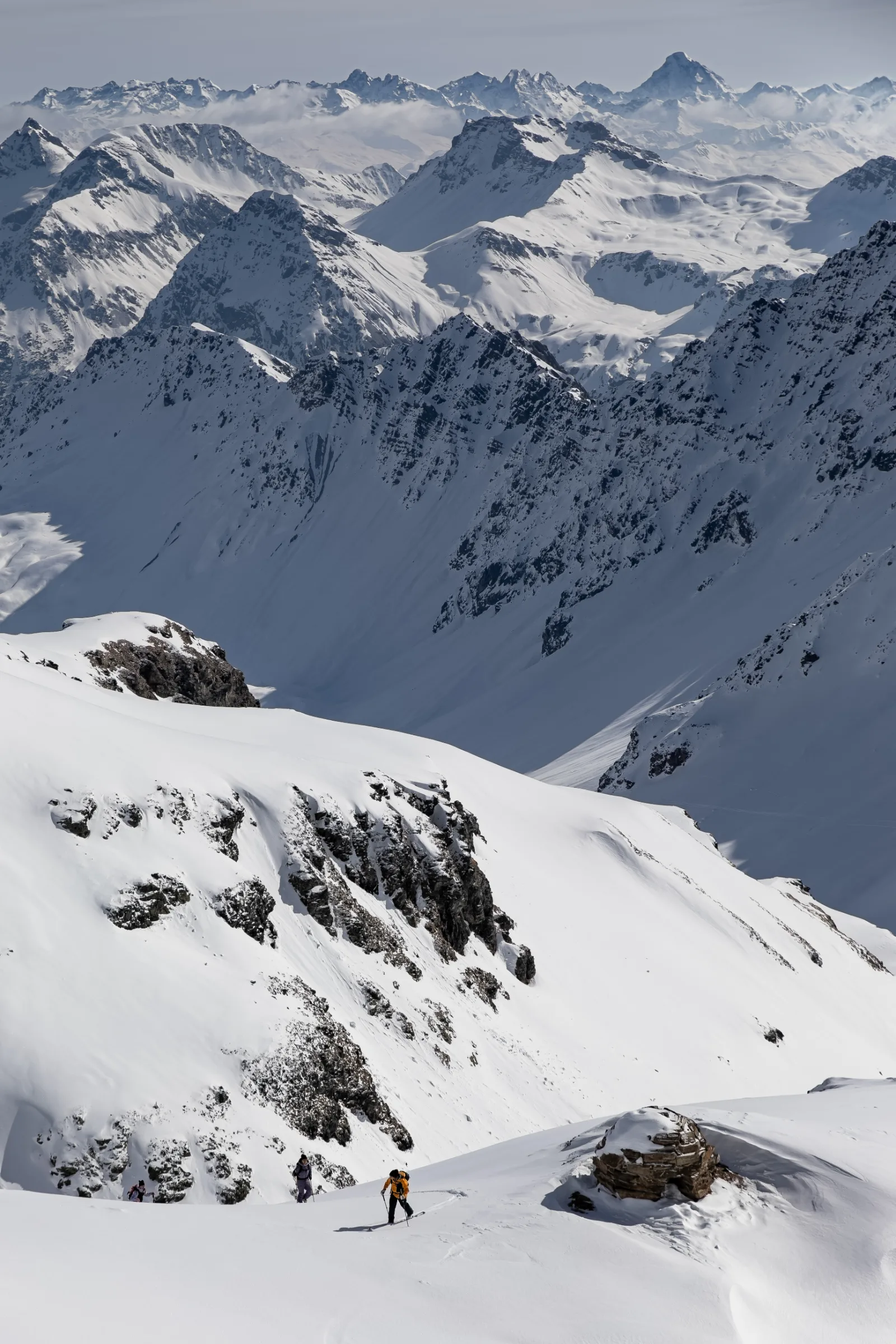 Drei Skitourengehende steigen bei Lenzerheide einen Schneehang hinauf, überragt von einem weiten Panorama schneebedeckter Felsgipfel.
