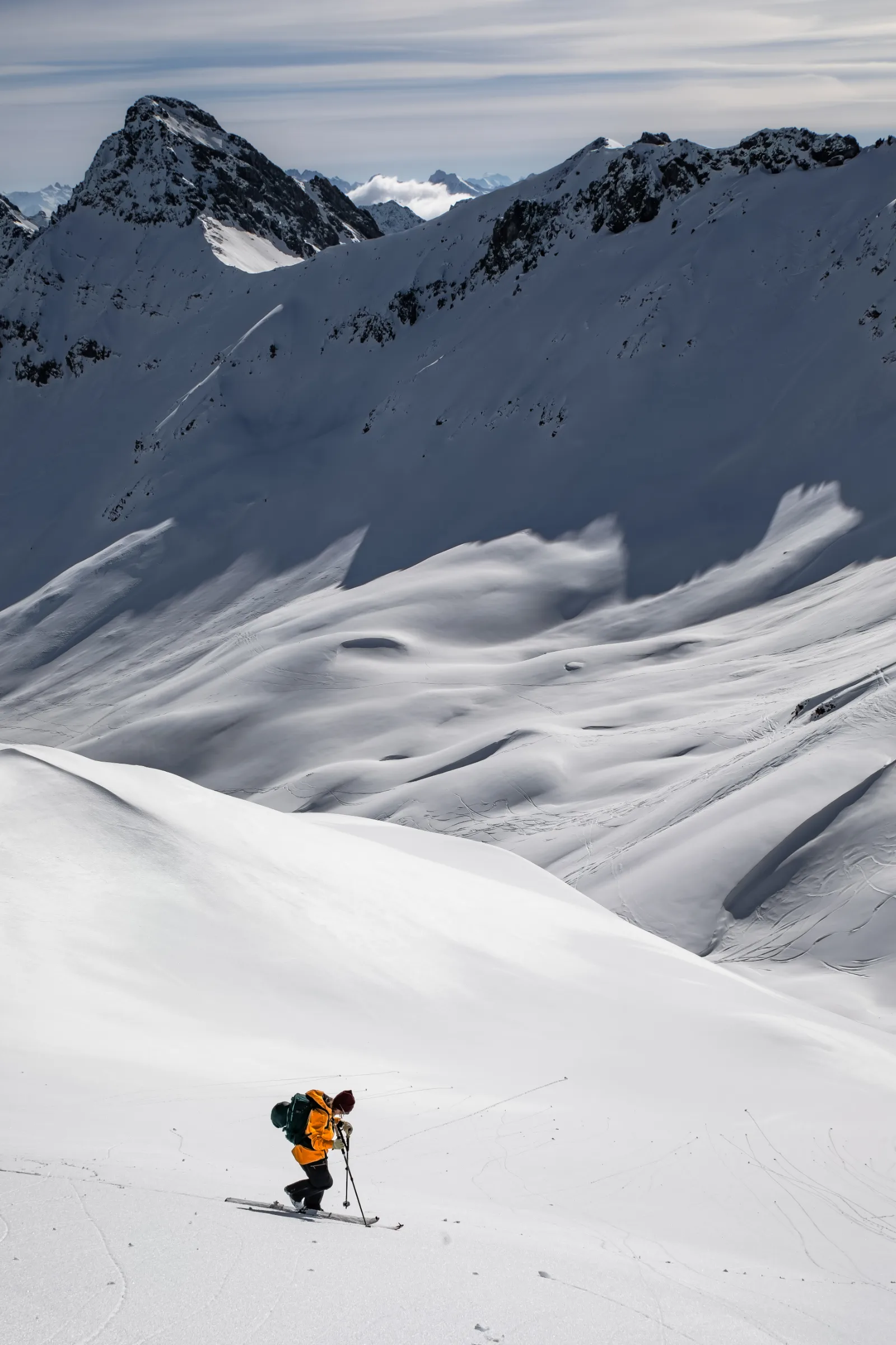 Michaela Casotti steigt in orangefarbener Jacke mit grünem Rucksack auf Tourenski bergauf, hinter ihr liegt ein weites Schneebecken mit schroffen Gipfeln.