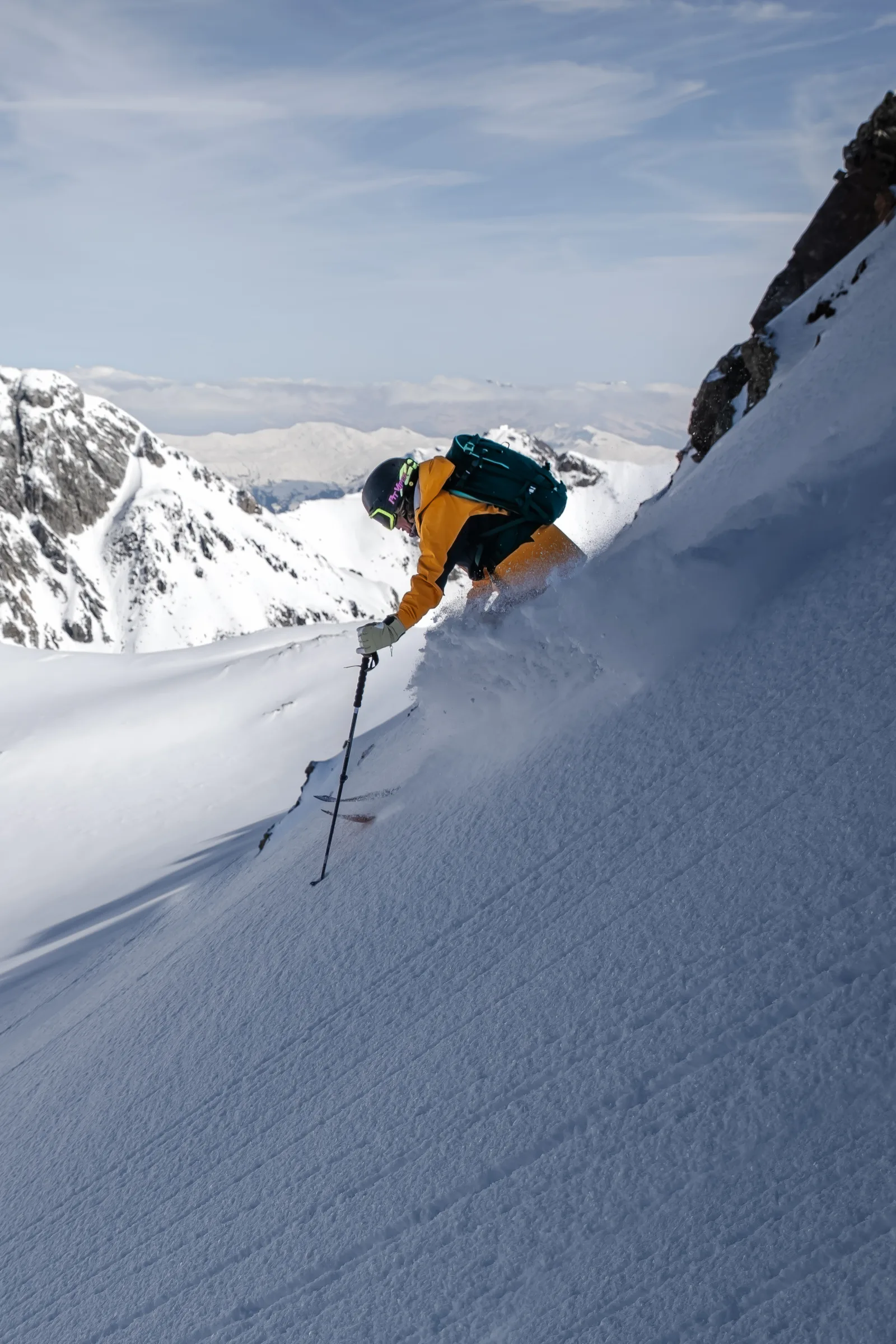 Michaela Casotti fährt tief gebeugt neben einer Felswand durch einen steilen Schneehang und wirbelt Schnee auf, mit verschneiten Gipfeln im Hintergrund.