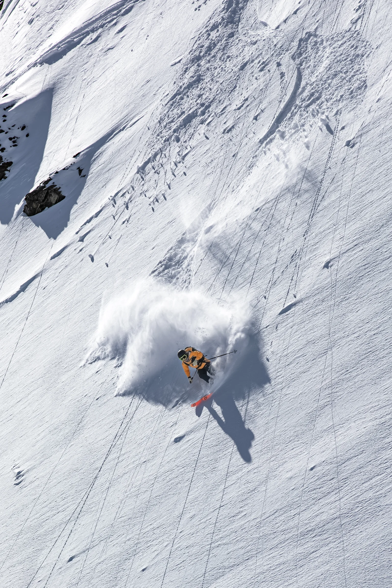 Michaela Casotti fährt einen Schwung auf einem sonnigen Steilhang und wirbelt eine große Schneewolke auf, während ihr langer Schatten talwärts fällt.