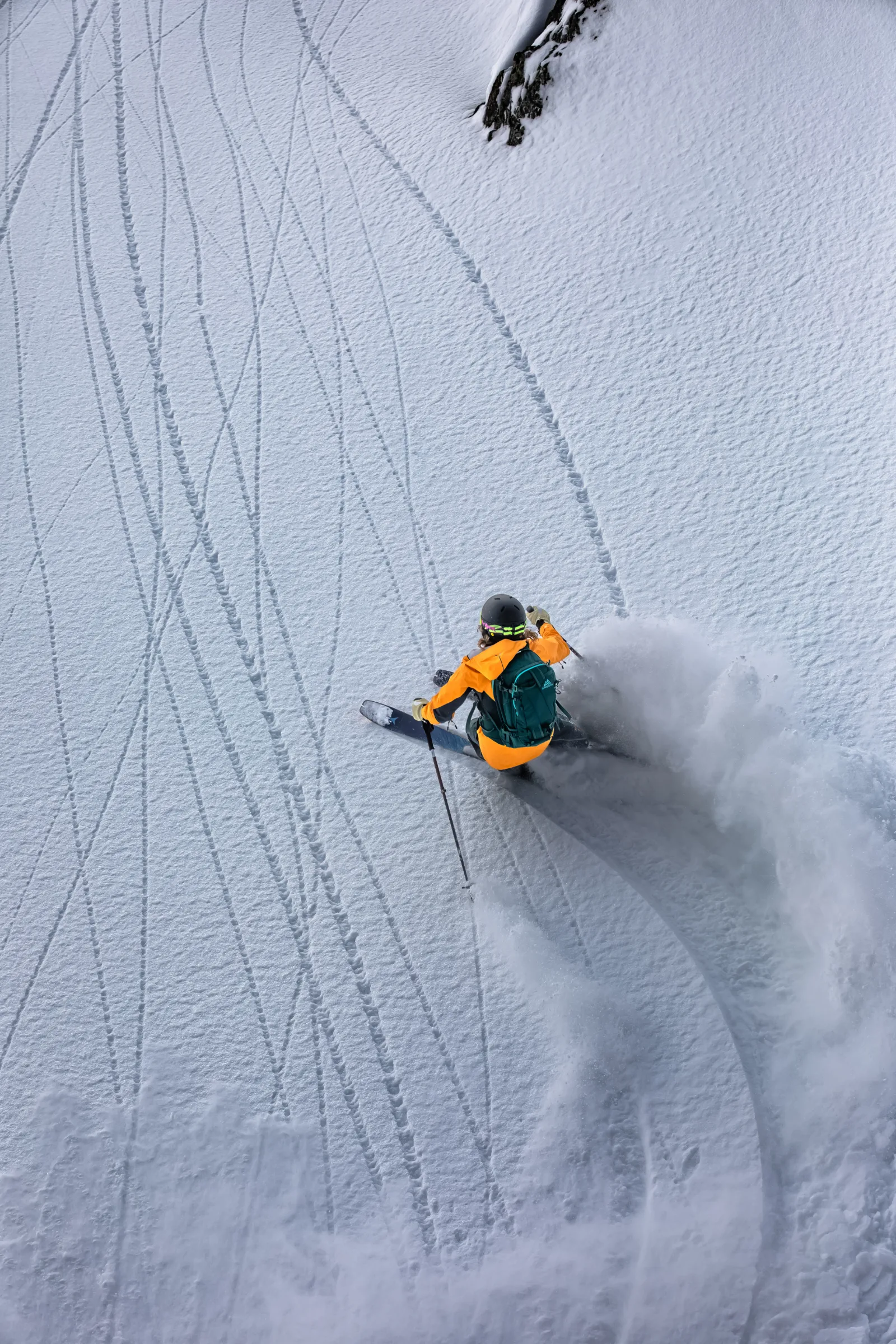Von hinten gesehen fährt Michaela Casotti mit orangefarbener Jacke und grünem Rucksack einen tiefen Schwung auf einem verspurten Schneehang und wirbelt Schnee auf.