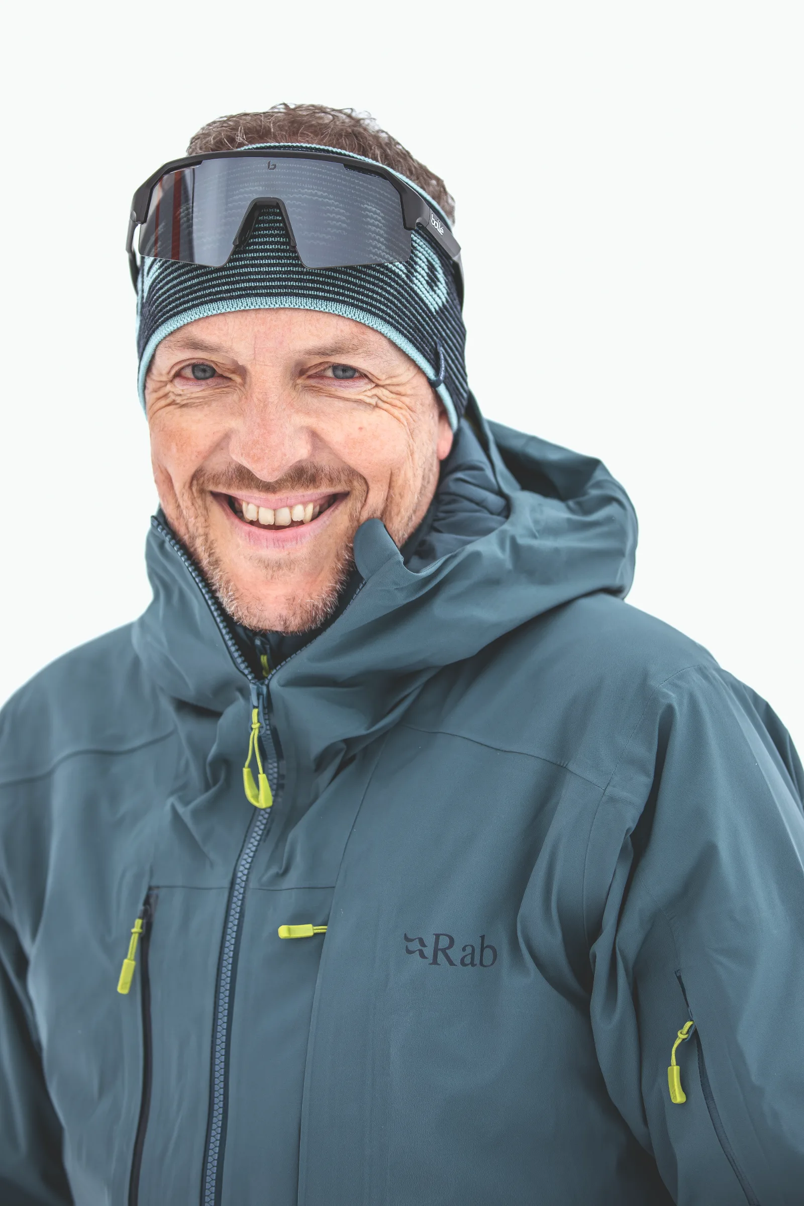 Mico Kehrle lächelt vor weißem Hintergrund in die Kamera, mit gestreiftem Stirnband, hochgeschobener Sonnenbrille und blaugrüner Jacke.