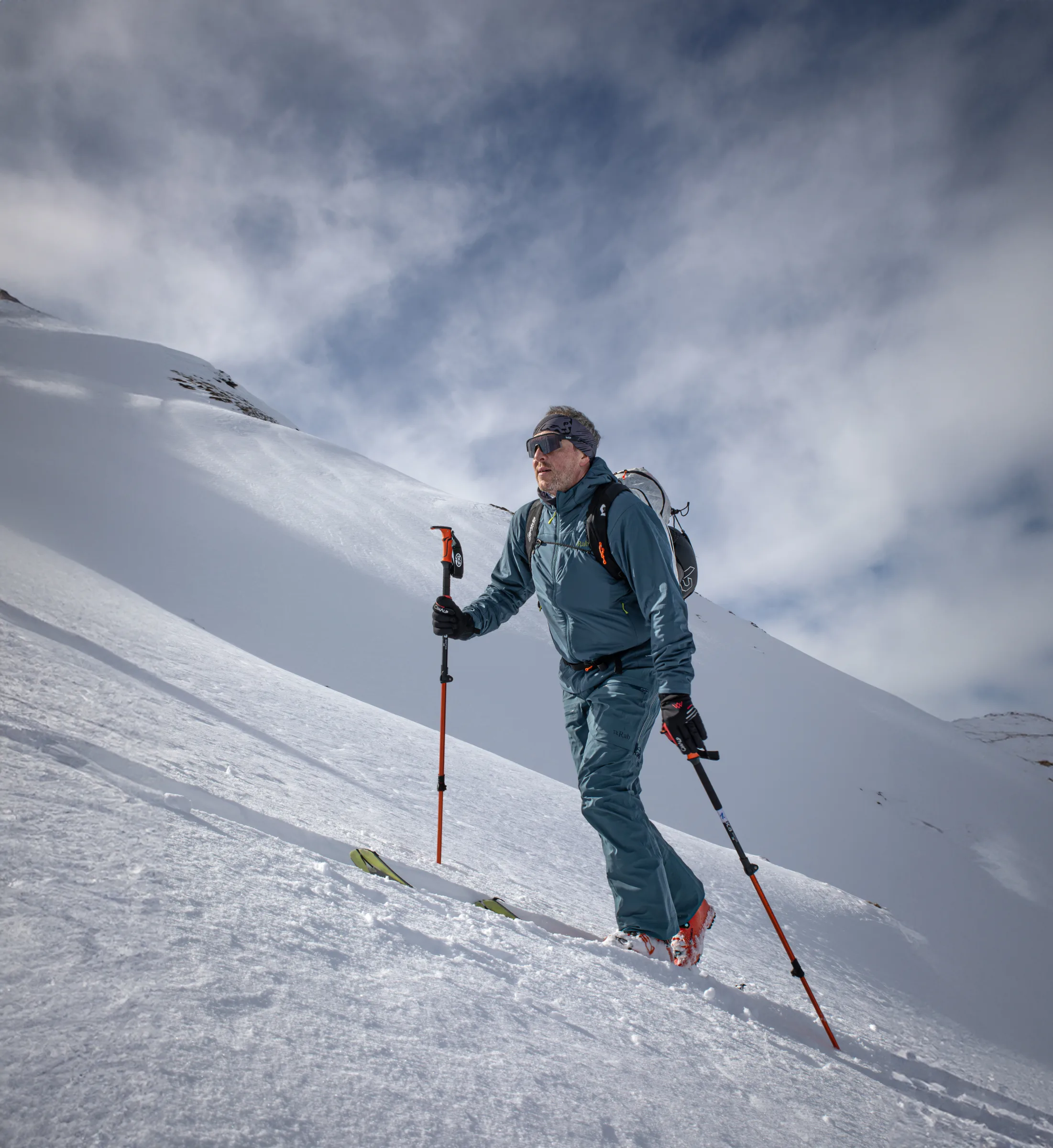 Mico Kehrle steigt mit Tourenski, Stöcken und Rucksack einen Schneehang in Gastein hinauf, hinter ihm verschneite Bergflanken und ein wolkiger Himmel.