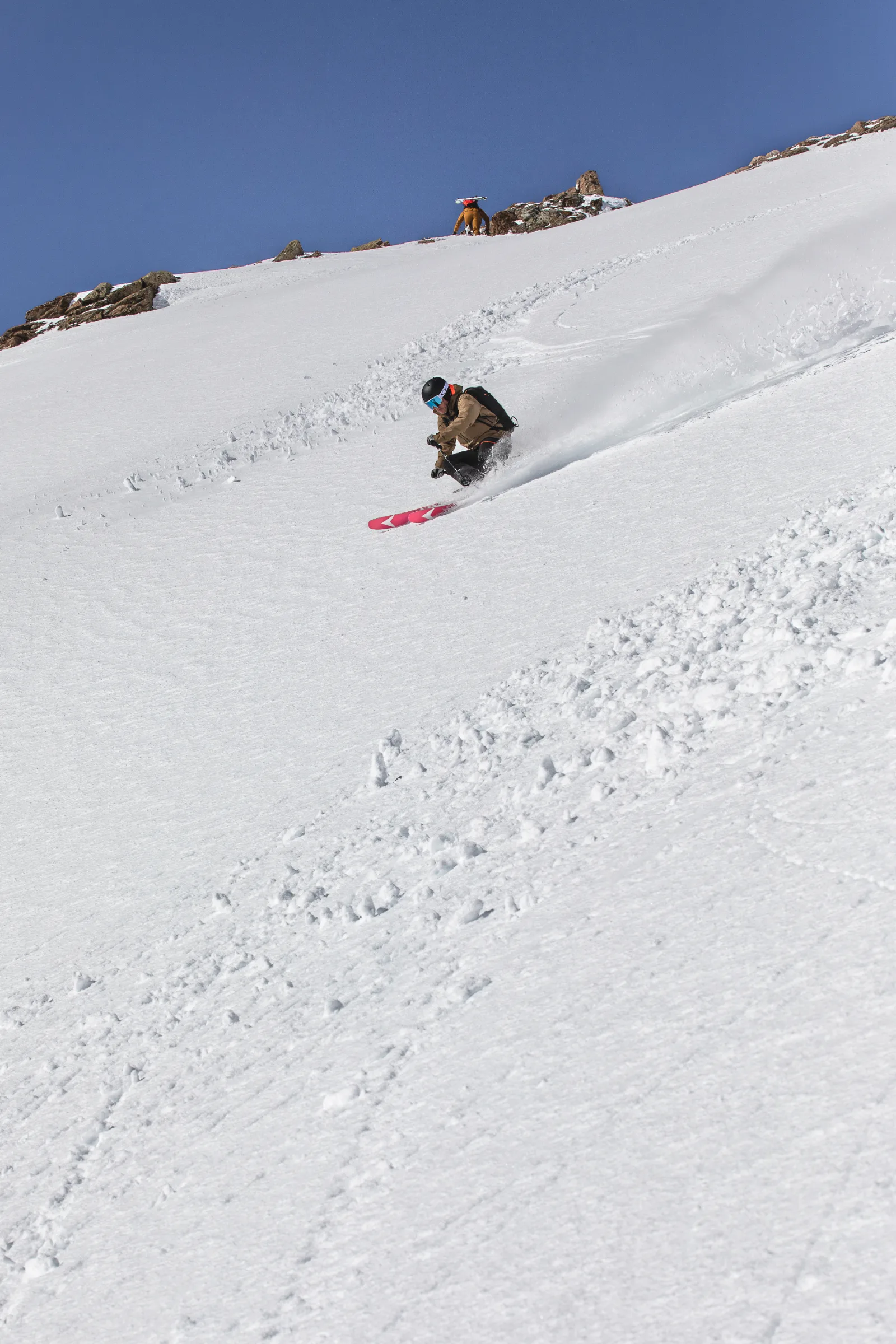 Mico Kehrle fährt auf roten Ski einen Schwung über einen steilen Schneehang, während weiter oben eine Person nahe dem felsigen Grat steht.