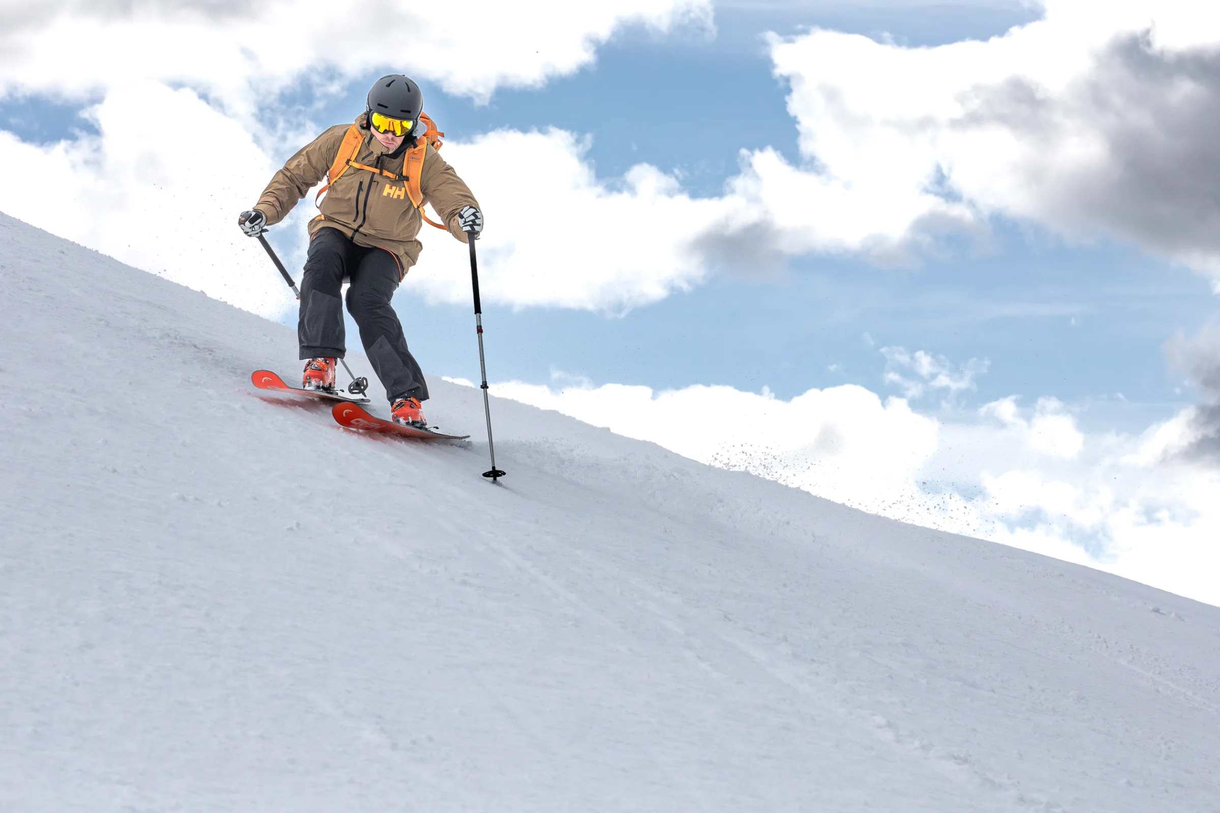 Mico Kehrle fährt mit gelb verspiegelter Skibrille und orangefarbenem Rucksack über einen Schneehang, aus tiefer Perspektive vor blauem Himmel und Wolken gesehen.