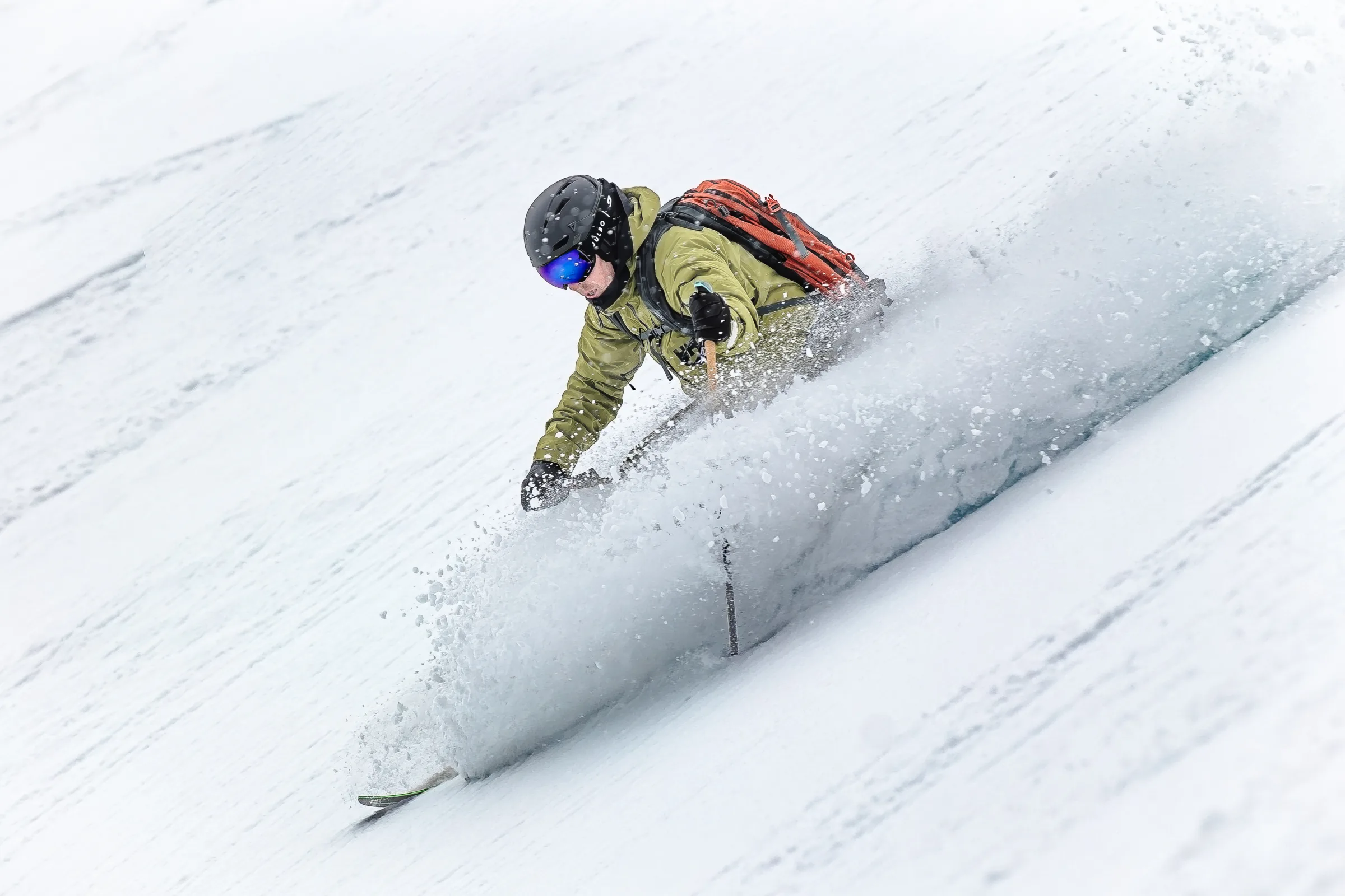 Mico Kehrle fährt tief gebeugt durch Pulverschnee, eine Hand dicht über der Schneeoberfläche, während eine breite Schneewolke seine Ski umhüllt.