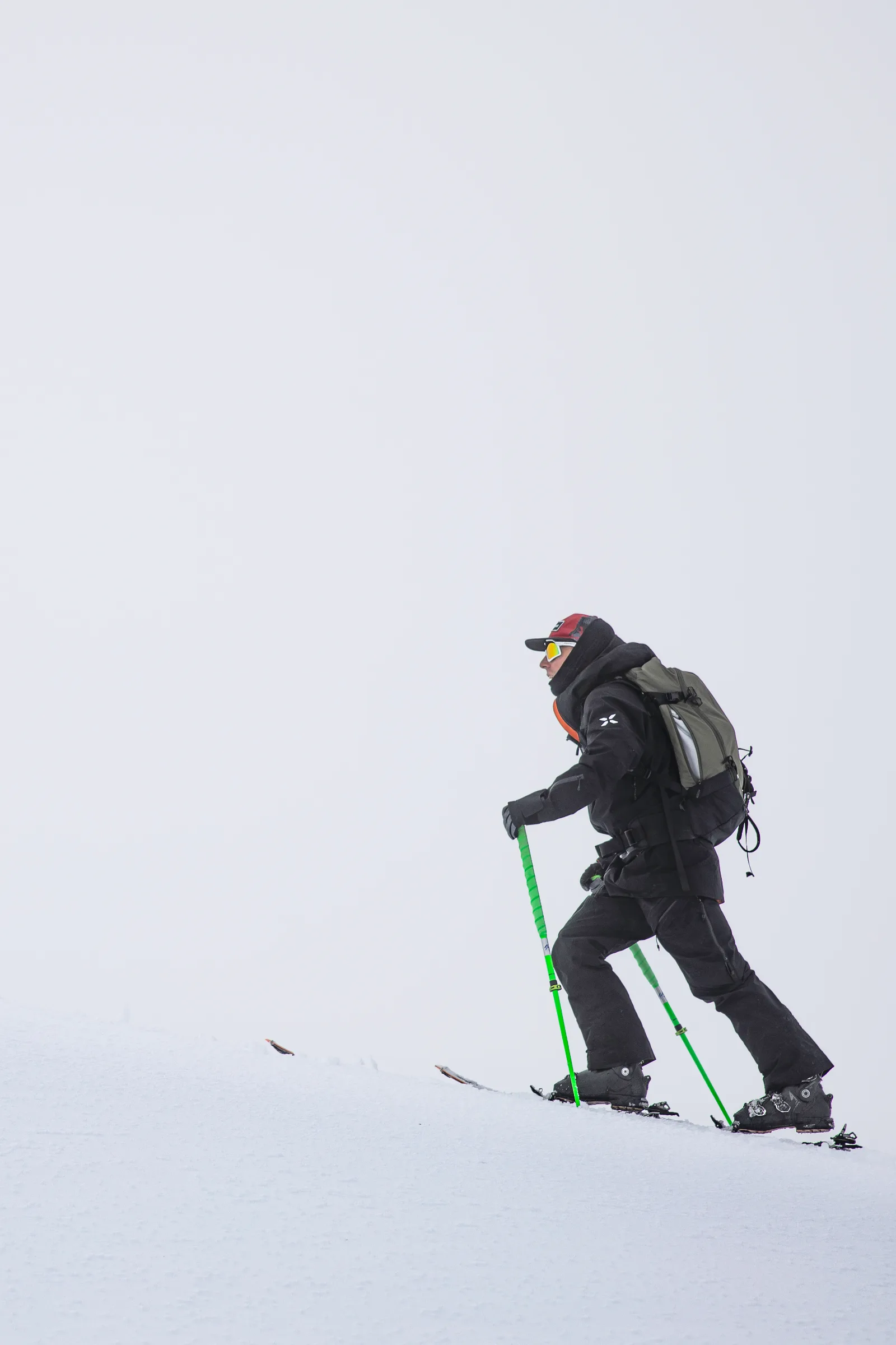 Mike Perl steigt mit Ski, grünen Stöcken und großem Rucksack einen Schneehang vor blassgrauem Himmel hinauf.