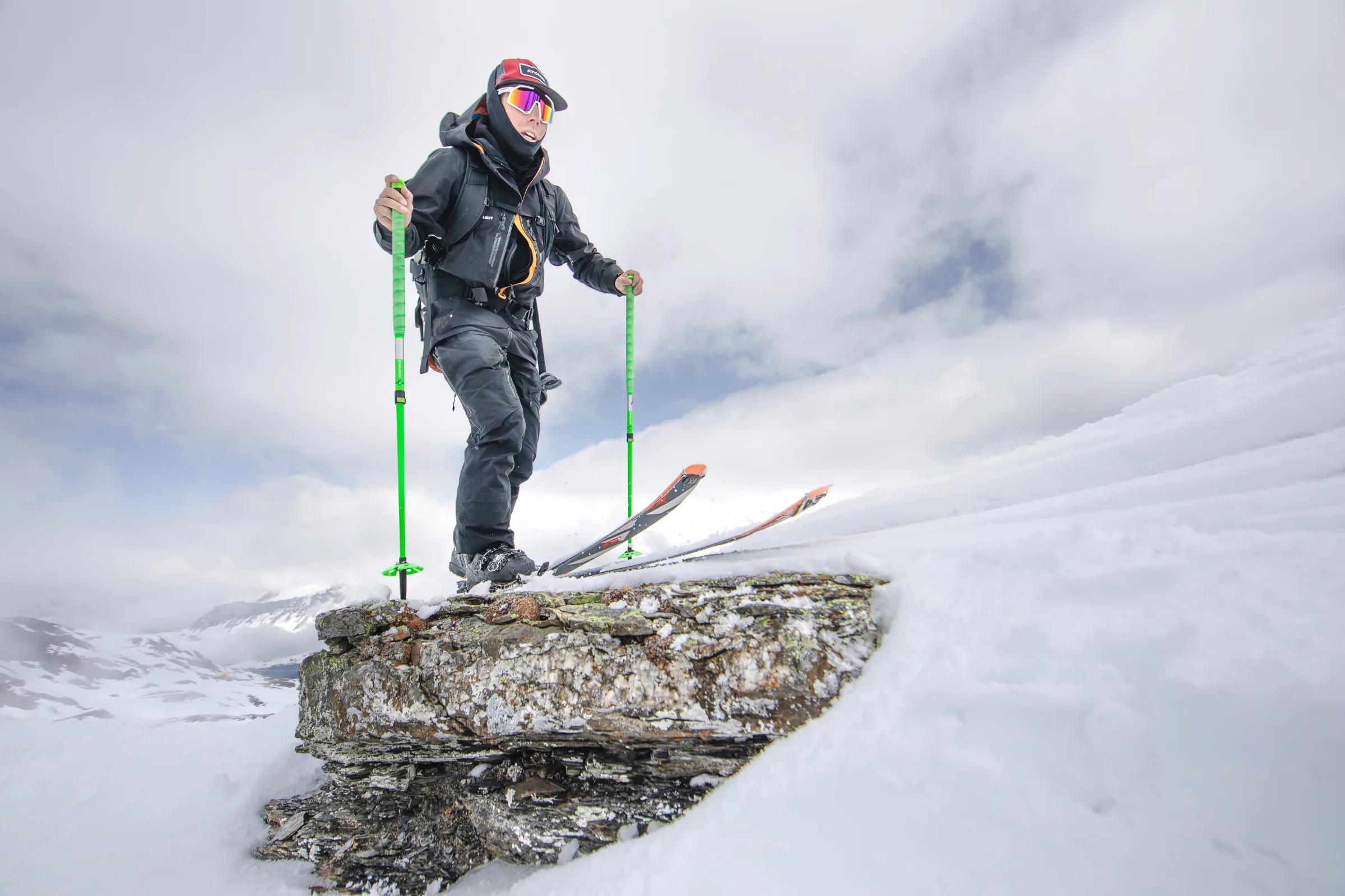 Mike Perl steht mit Ski und grünen Stöcken auf einer schneebedeckten Felskante, hinter ihm liegen Berge unter tiefen Wolken.