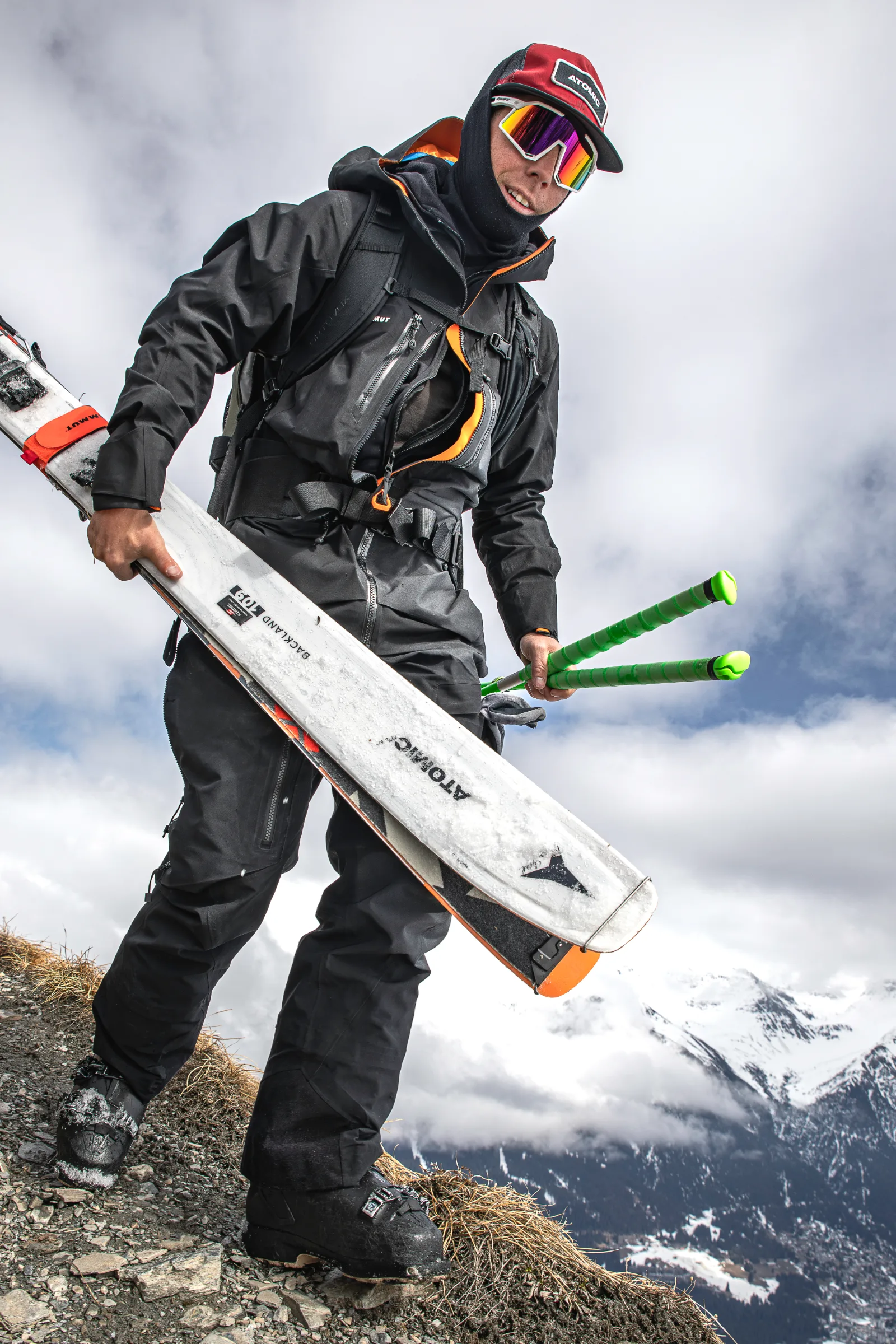 Aus tiefer Perspektive ist Mike Perl mit Ski und grünen Stöcken auf einem steinigen Grat zu sehen, dahinter wolkenverhangene Schneeberge.