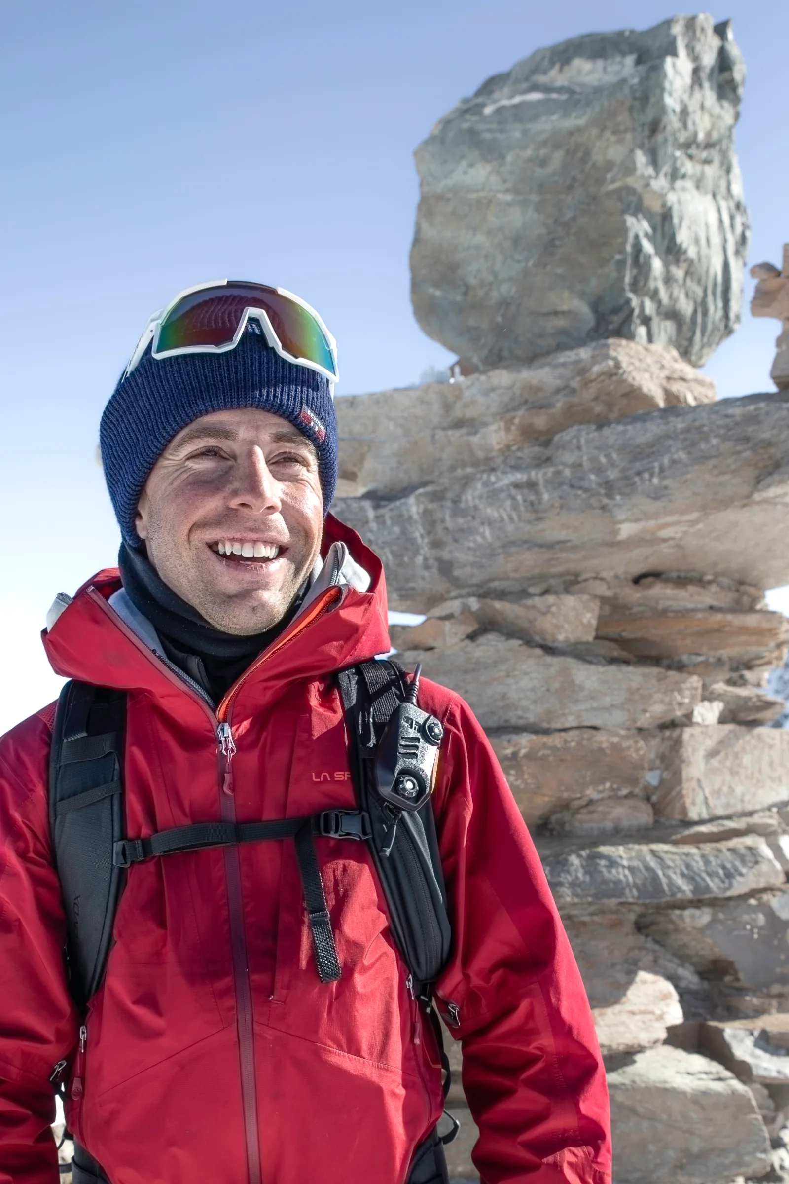 Mike Perl lächelt in roter Jacke und blauer Mütze vor aufgeschichteten Felsplatten, die Sonnenbrille sitzt auf seiner Mütze.