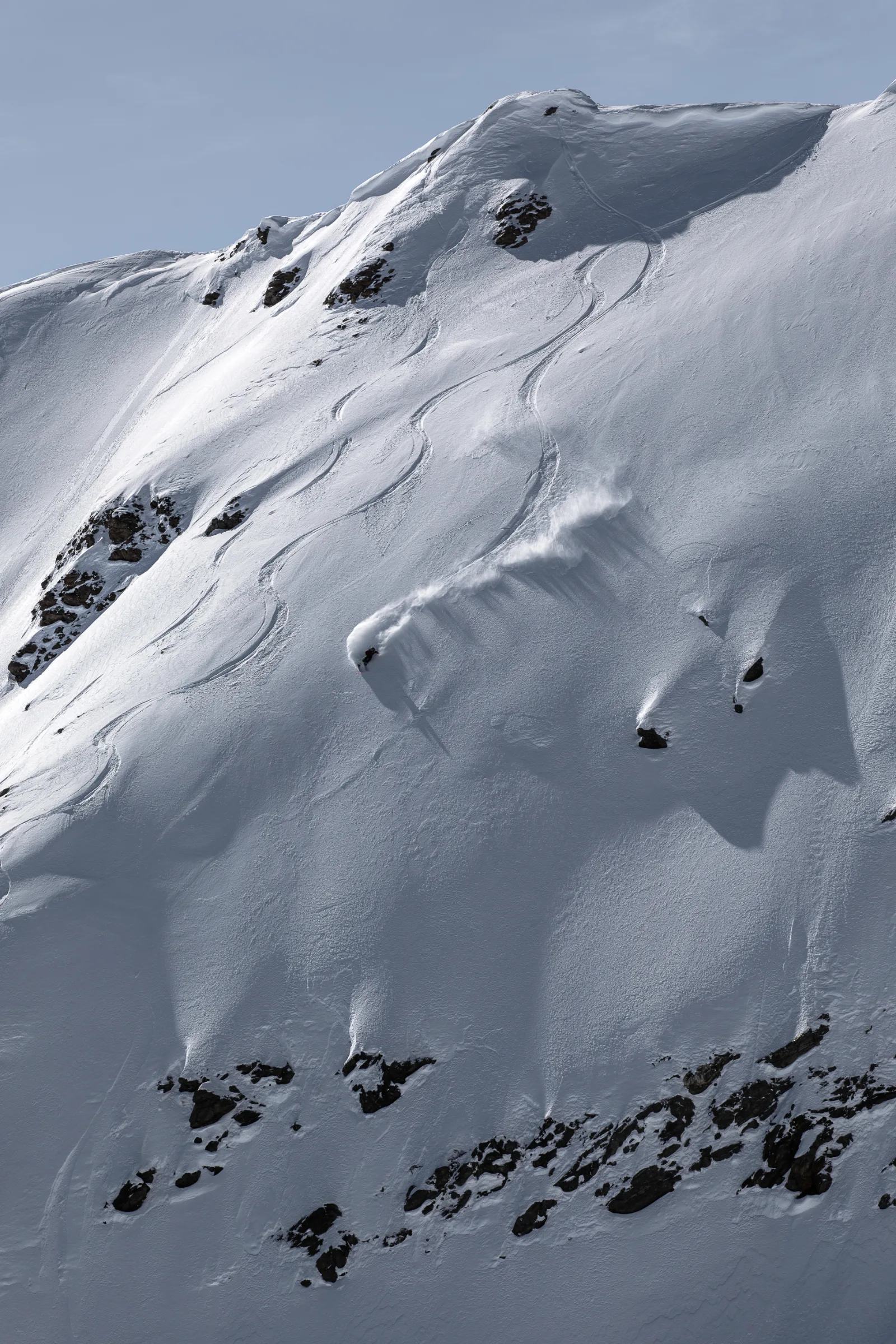 Mike Perl fährt einen weiten Schwung im Pulverschnee, eine lange Schneefahne und geschwungene Spuren ziehen sich über den felsdurchsetzten Hang.