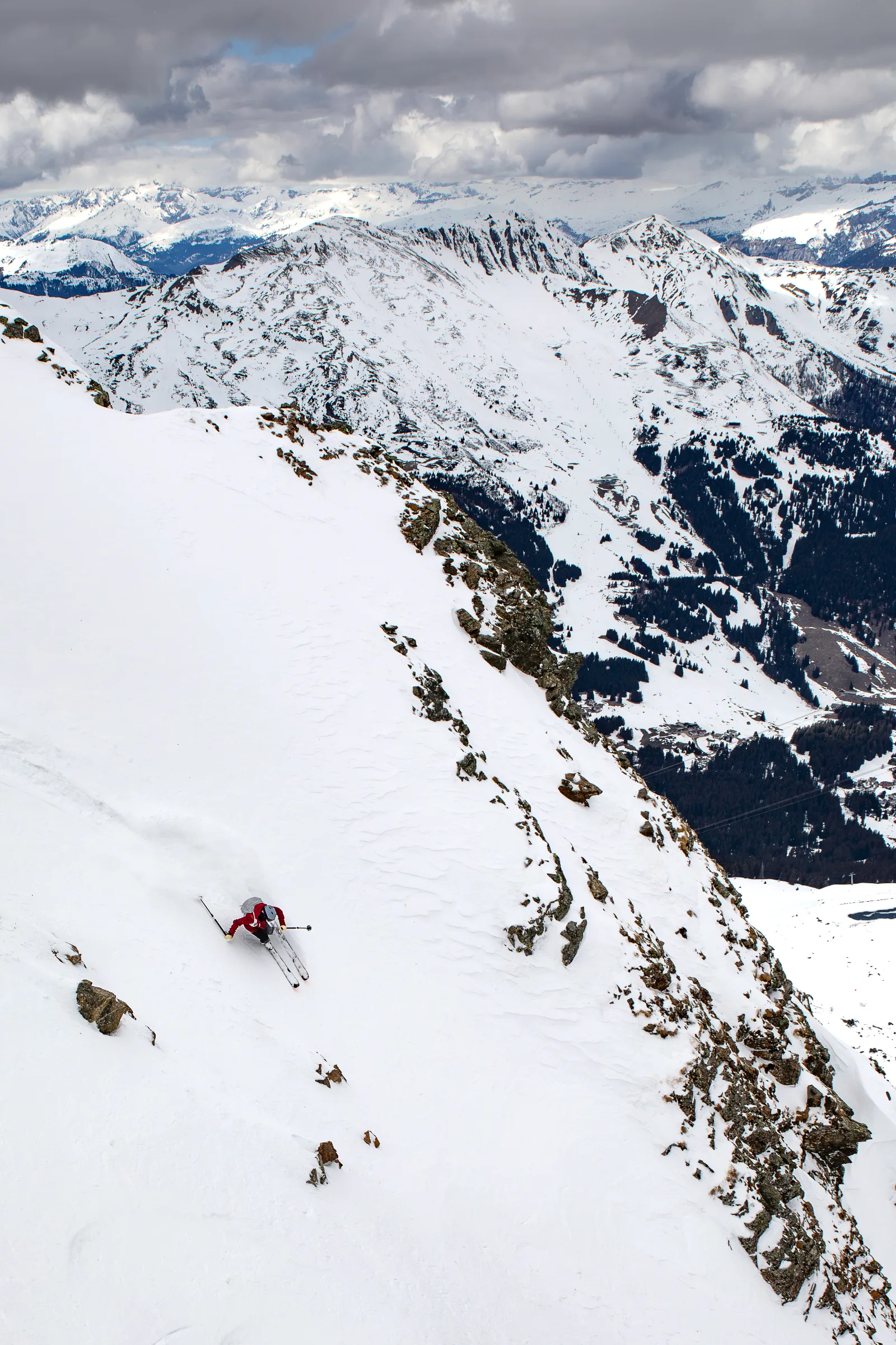 Mike Perl fährt in roter Jacke einen steilen Schneehang zwischen Felsen hinab, vor einem weiten Panorama verschneiter Berge und bewaldeter Täler unter dichten Wolken.