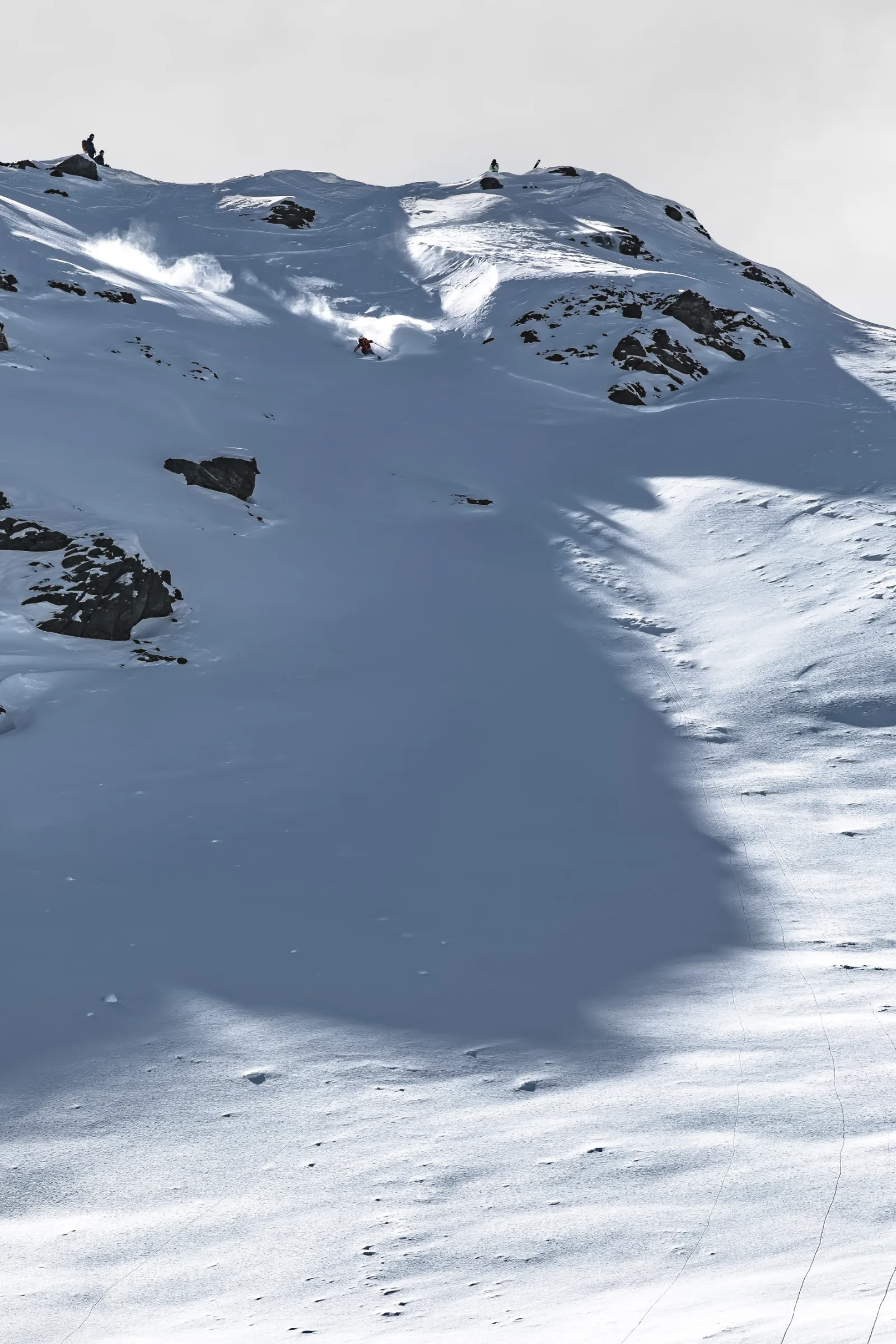 Mike Perl zieht einen Schwung durch einen steilen, felsdurchsetzten SchneeHang und wirbelt eine Schneefahne auf, während oben Menschen am Grat stehen.