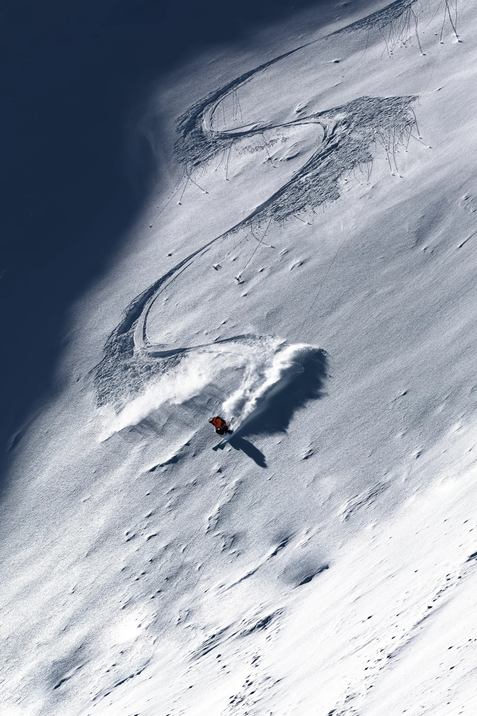 Mike Perl fährt in orangefarbener Jacke über einen steilen Schneehang und hinterlässt geschwungene Spuren sowie eine breite Pulverschneefahne.