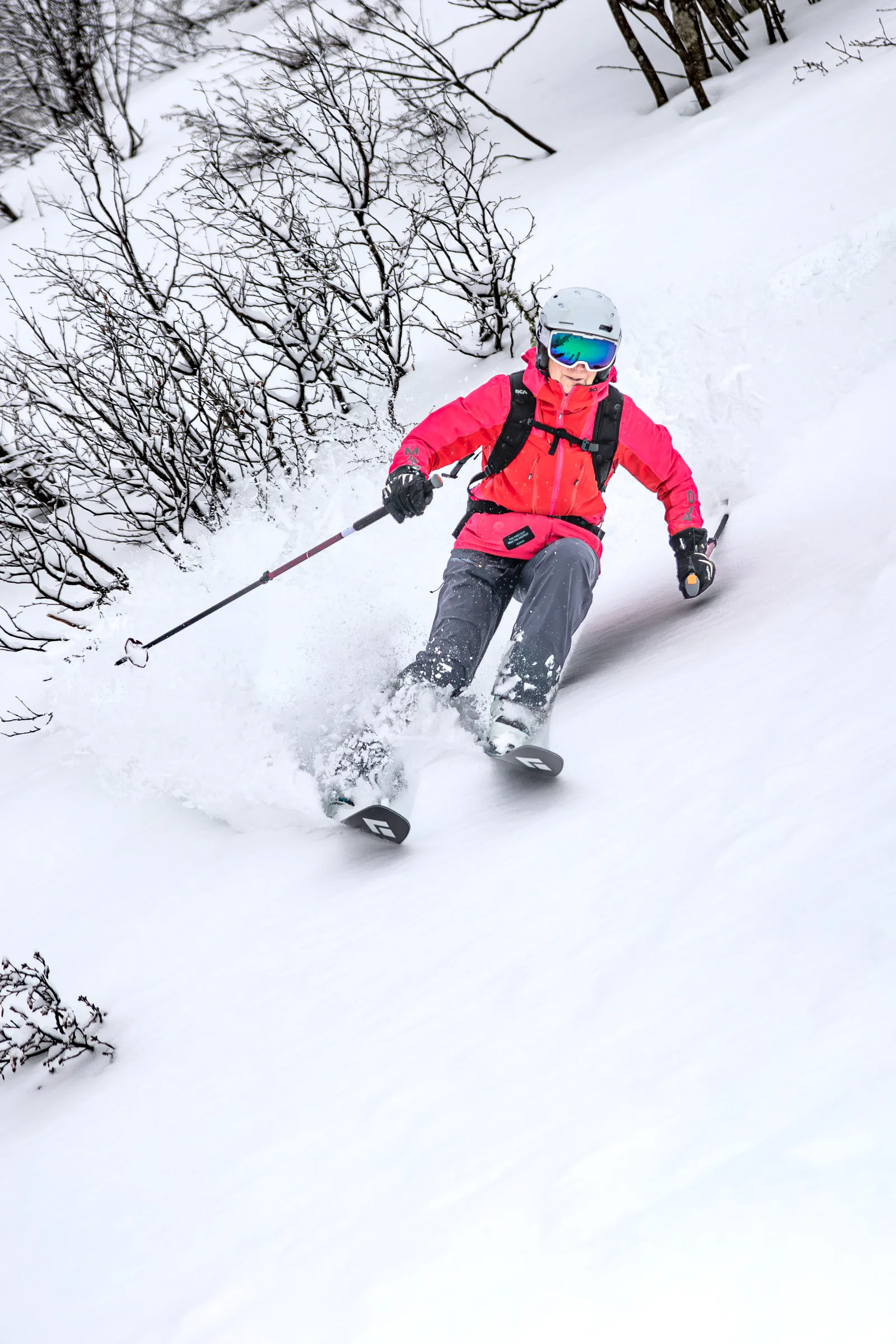 Petra Rapp fährt in pink-roter Jacke mit Helm und Rucksack durch tiefen Schnee zwischen kahlen Bäumen in Gastein und wirbelt Schnee auf.