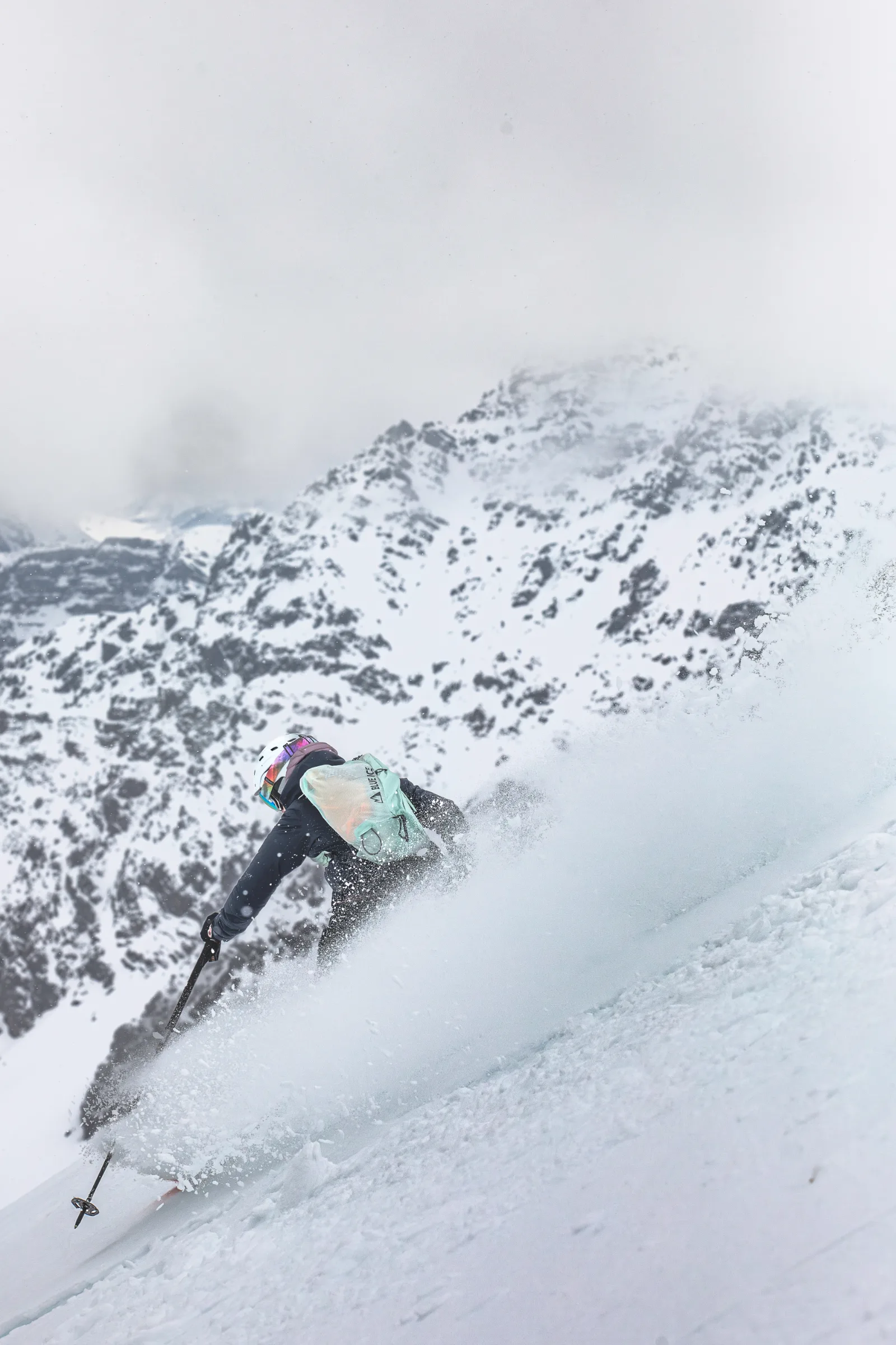 Von hinten gesehen zieht Petra Rapp einen Schwung durch einen steilen Schneehang und wirbelt eine große Schneewolke auf, vor felsigen, verschneiten Bergen.