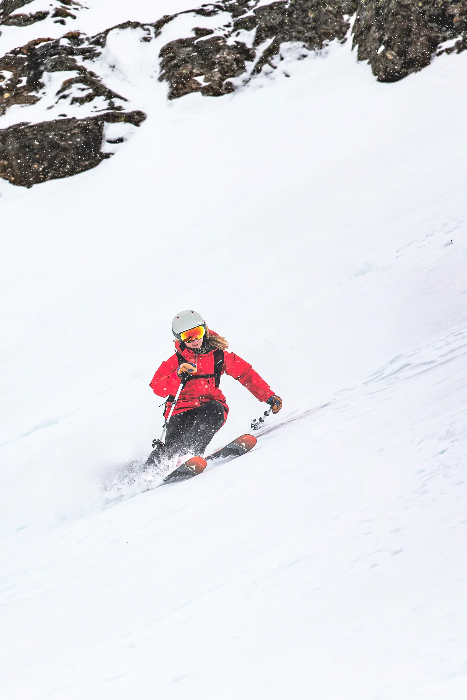 Pia Vogt fährt mit roter Jacke, grauem Helm und verspiegelter Brille einen Schwung auf einem Schneehang unter dunklen Felsen.