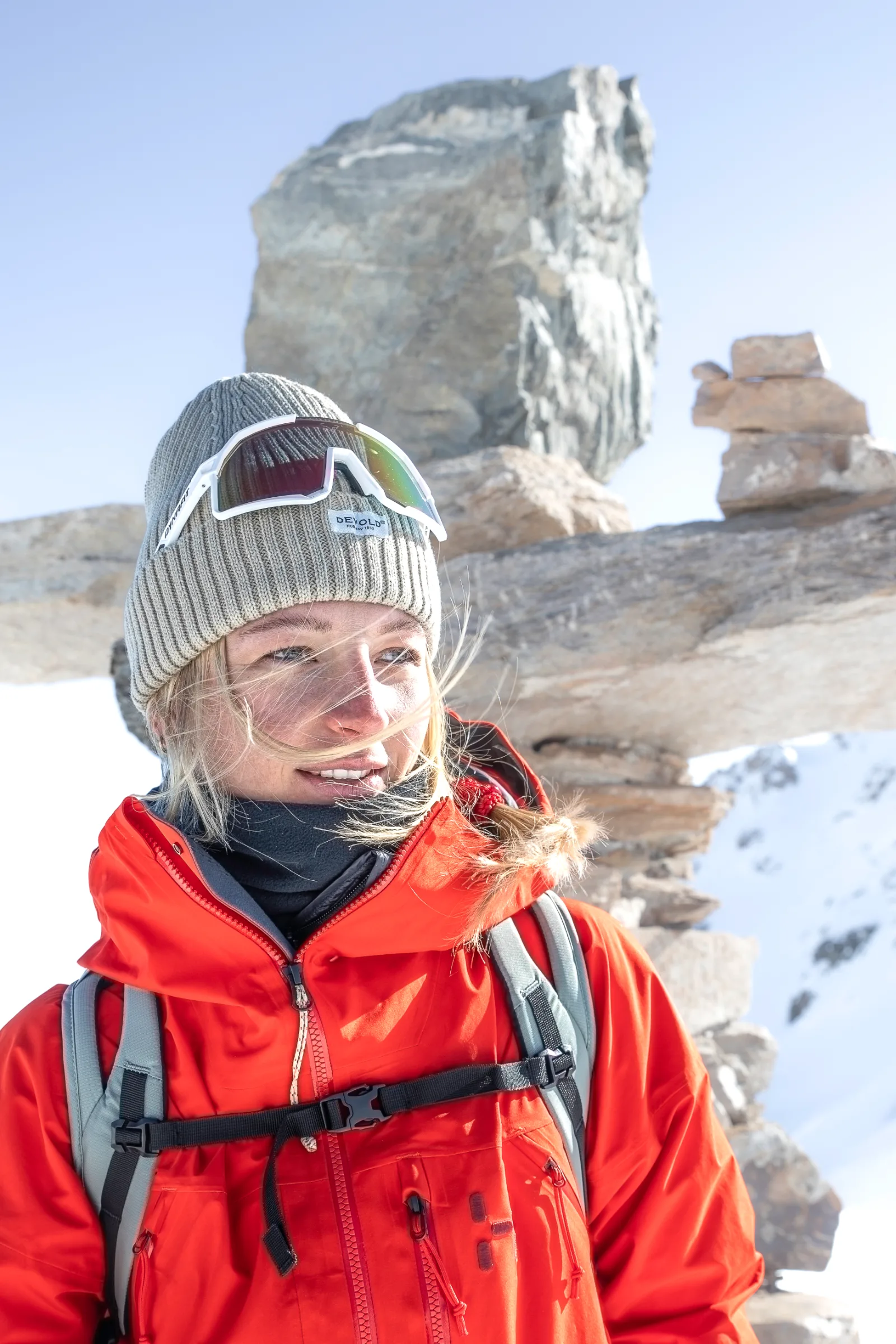 Pia Vogt blickt lächelnd zur Seite, mit grauer Strickmütze und darauf sitzender Sonnenbrille, vor Felsblöcken und Schnee unter blauem Himmel.