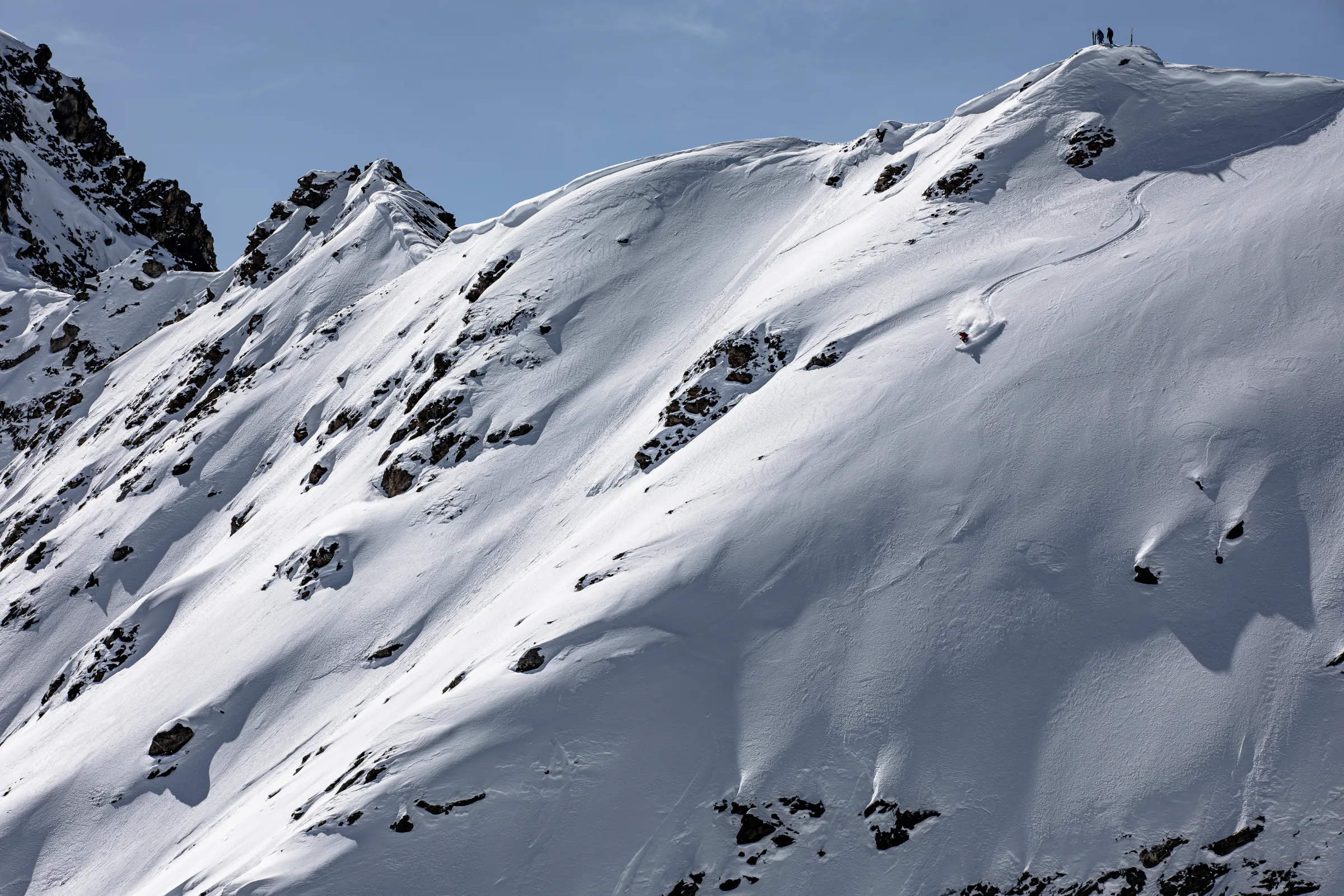 Pia Vogt zieht auf einem steilen, felsdurchsetzten Schneehang in Lenzerheide einen Schwung mit Schneefahne, während mehrere Personen oben auf dem Grat stehen.