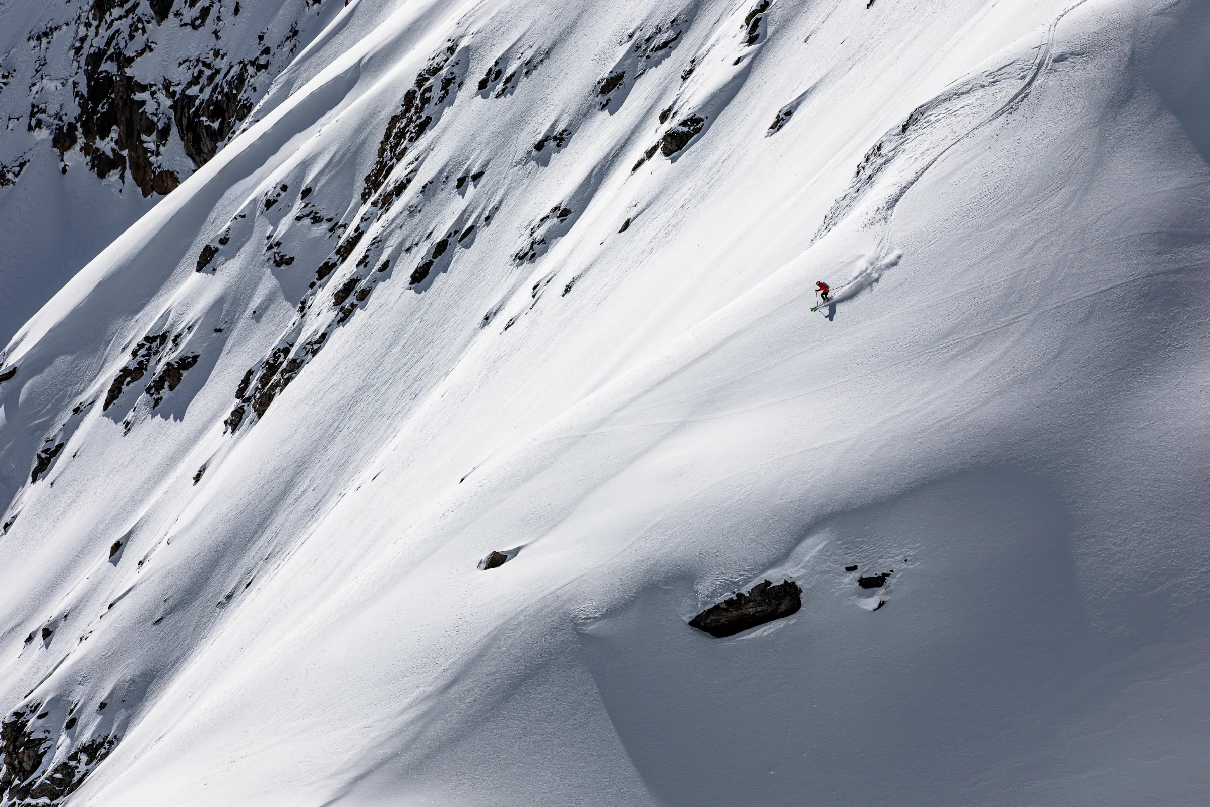 Pia Vogt fährt mit grünen Ski und roter Jacke über einen offenen Schneehang unterhalb felsiger Steilflanken und wirbelt dabei Schnee auf.