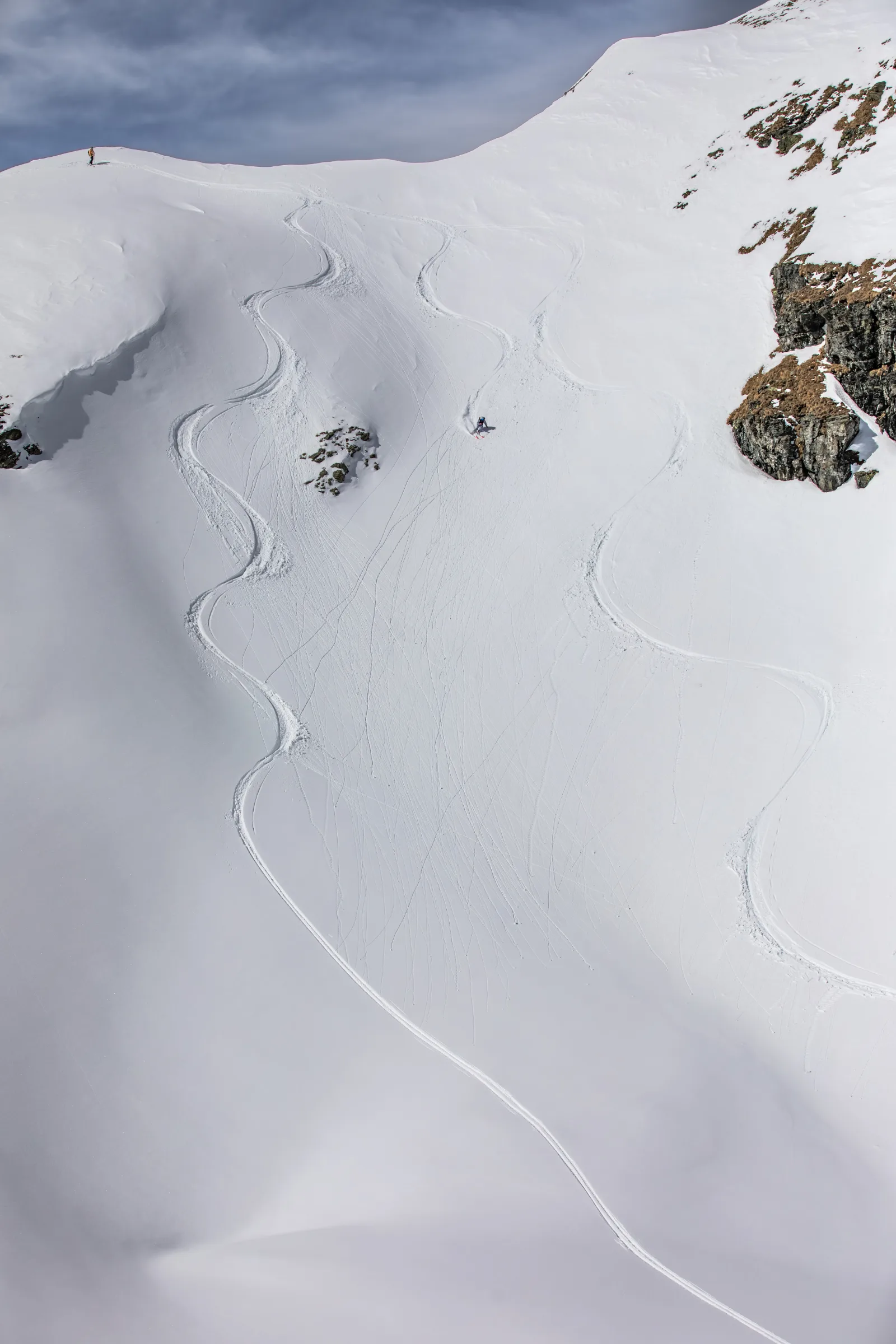 Pia Vogt fährt auf Ski einen weiten, felsgesäumten Schneehang in Lenzerheide hinab, während eine weitere Person oberhalb der geschwungenen Spuren am Grat steht.