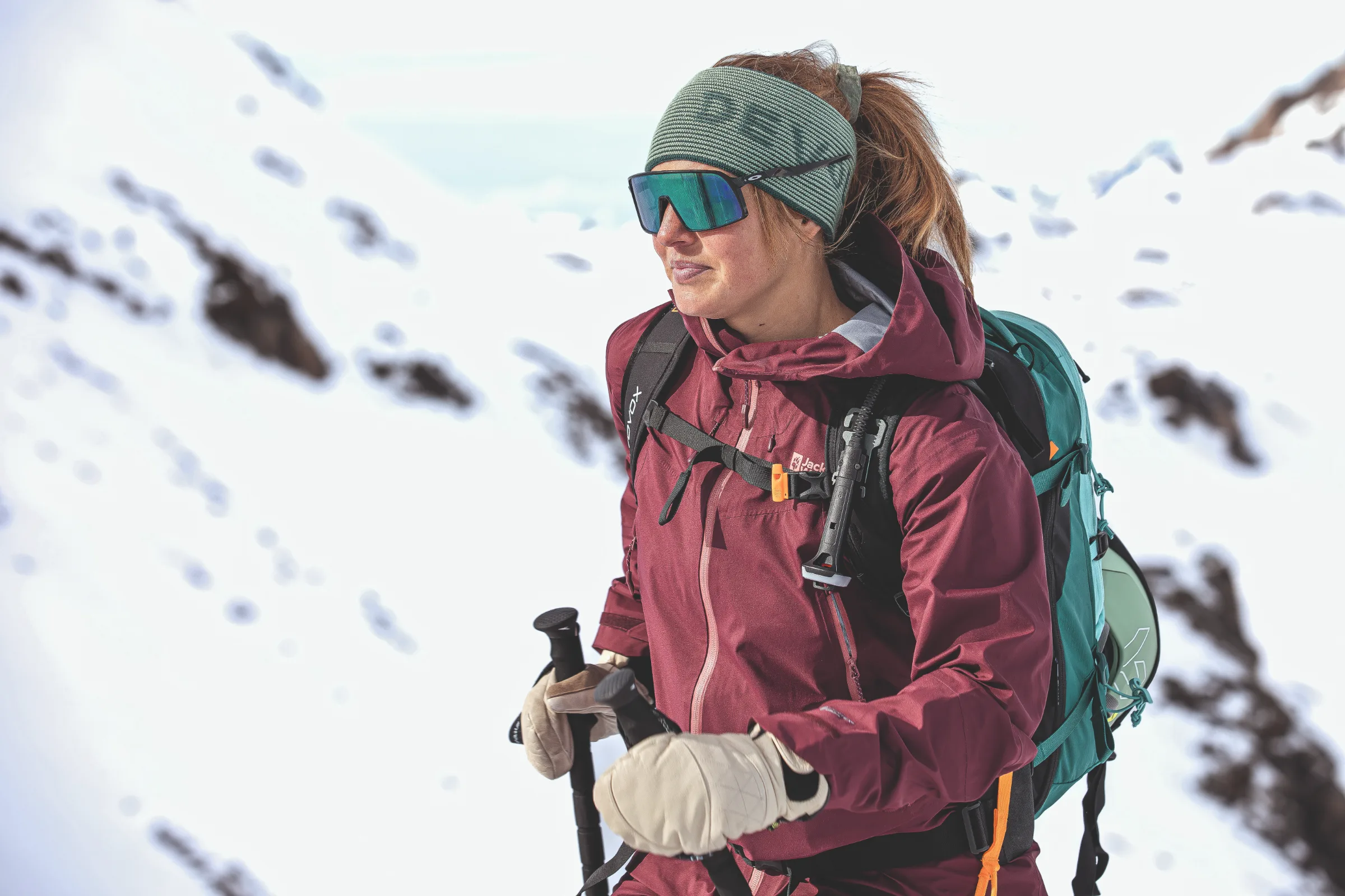 Ramona Arendt hält Skistöcke vor einem verschneiten Felshang und blickt zur Seite, mit grünem Stirnband, verspiegelter Sonnenbrille und türkisfarbenem Rucksack.