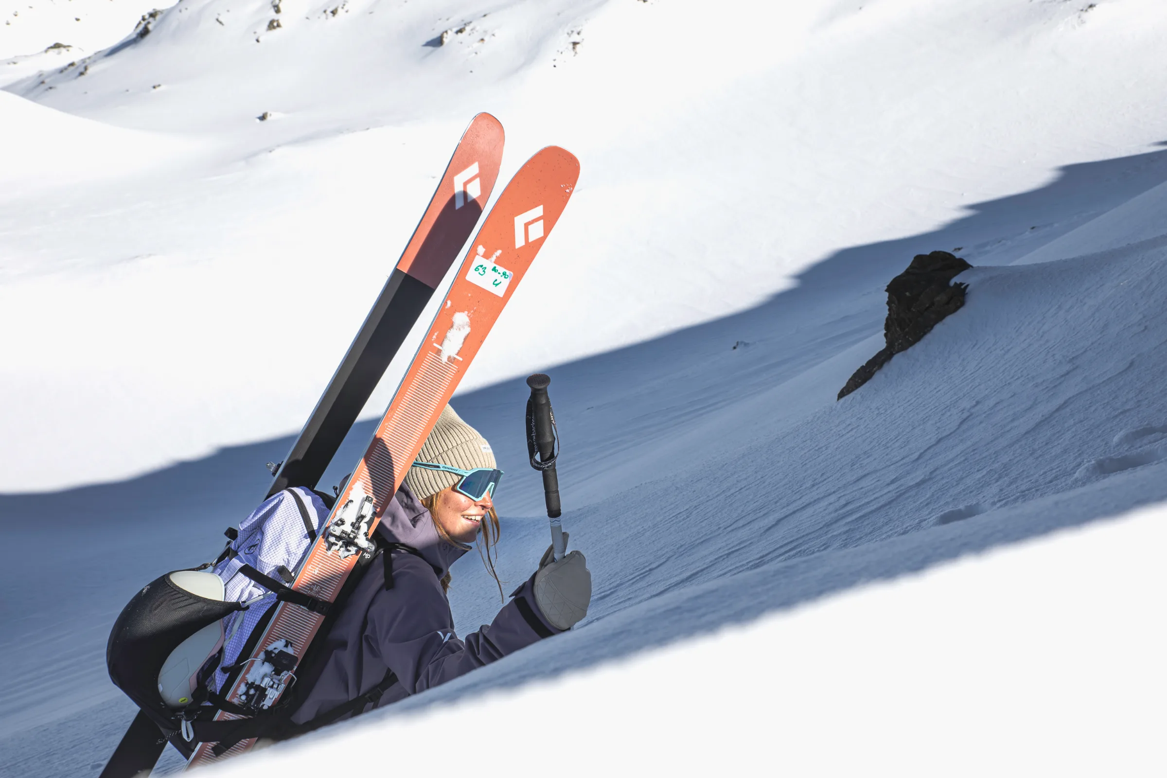 Ramona Arendt steigt lächelnd einen Schneehang hinauf, mit roten Ski und Helm am Rucksack und einem Stock in der Hand; hinter ihr ragt ein Felsen aus dem Schnee.