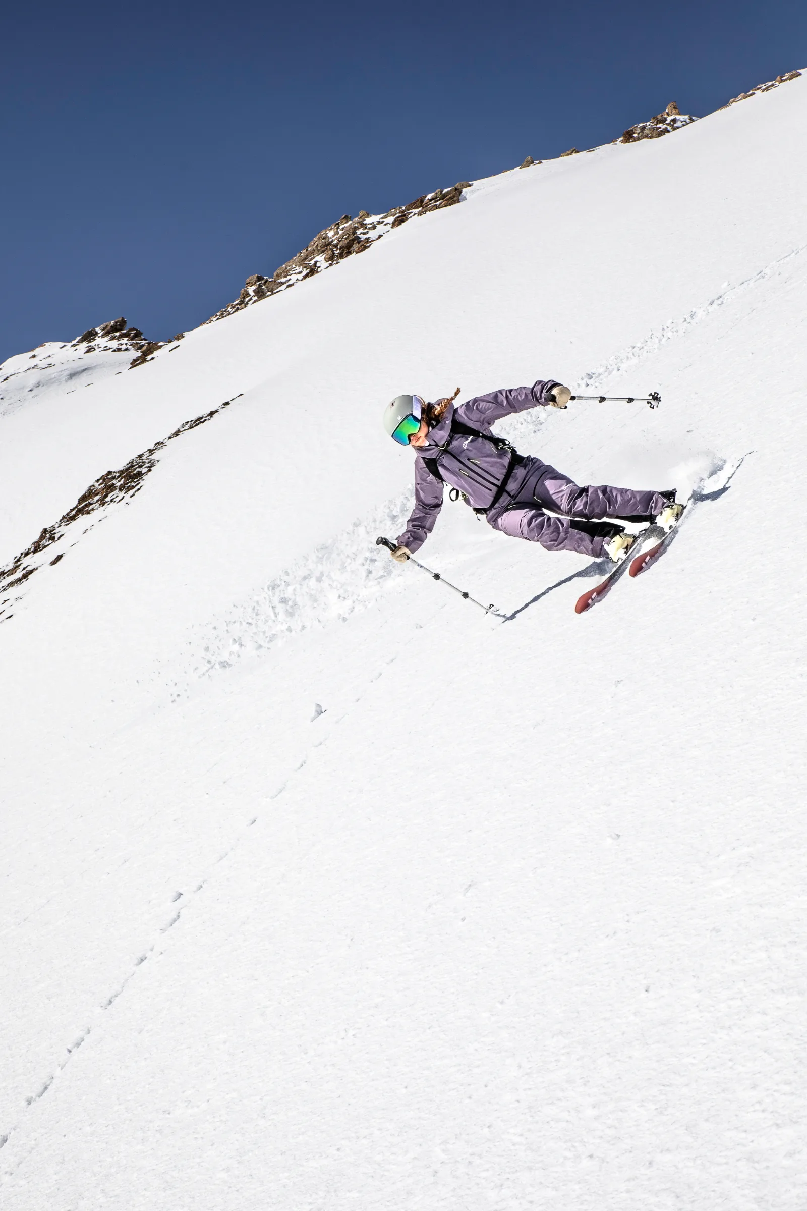 Ramona Arendt legt sich in violetter Skikleidung mit Helm und Skibrille in eine Kurve auf einem steilen Schneehang unter felsigem Grat und blauem Himmel.