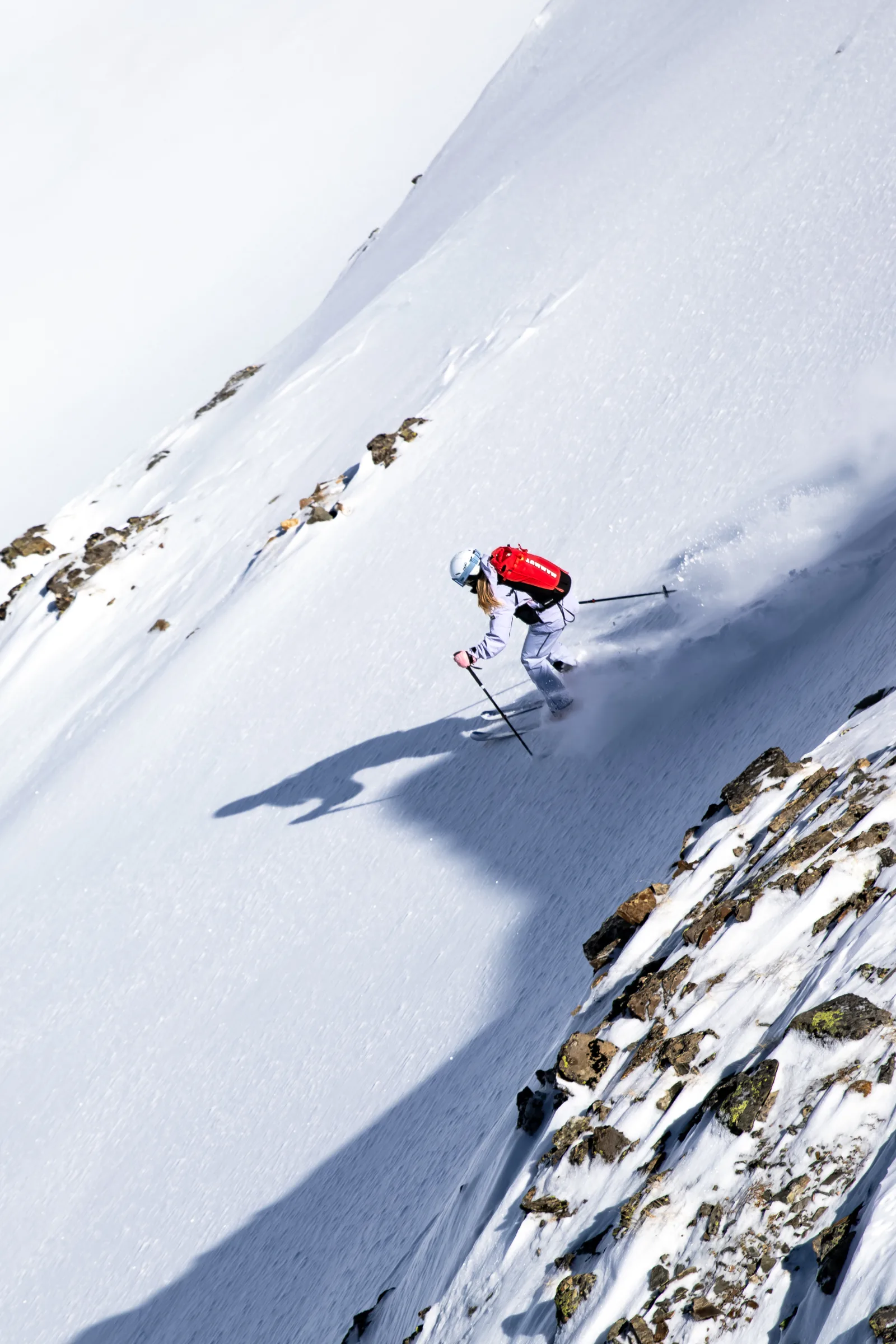 Ramona Arendt fährt in hellem Skianzug und mit rotem Rucksack neben einem felsigen Hangabschnitt durch den Schnee, der hinter ihren Ski aufwirbelt.