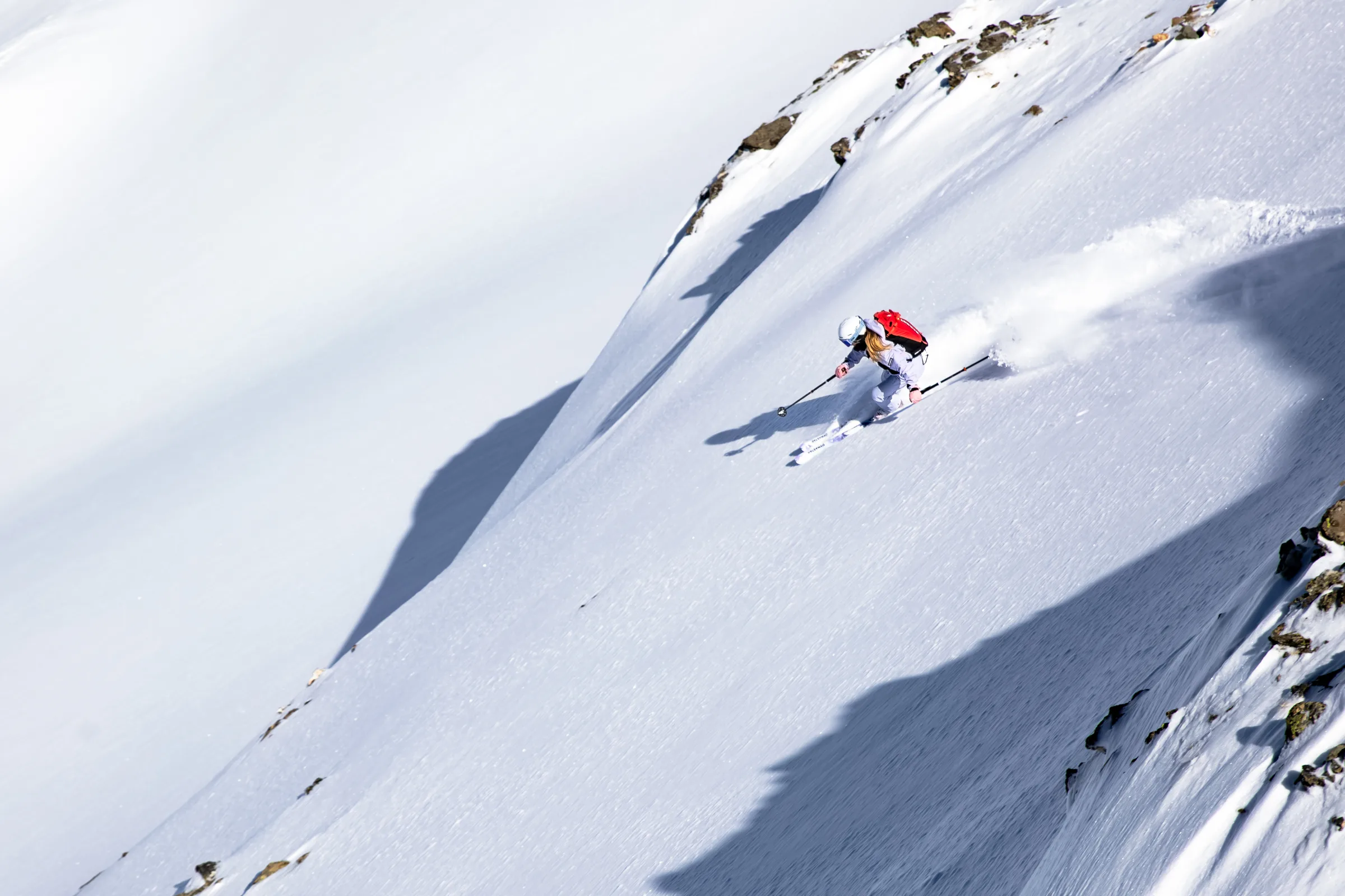 Ramona Arendt legt sich auf einem steilen, sonnigen Schneehang in die Kurve und wirbelt unterhalb freiliegender Felsen Schnee auf.