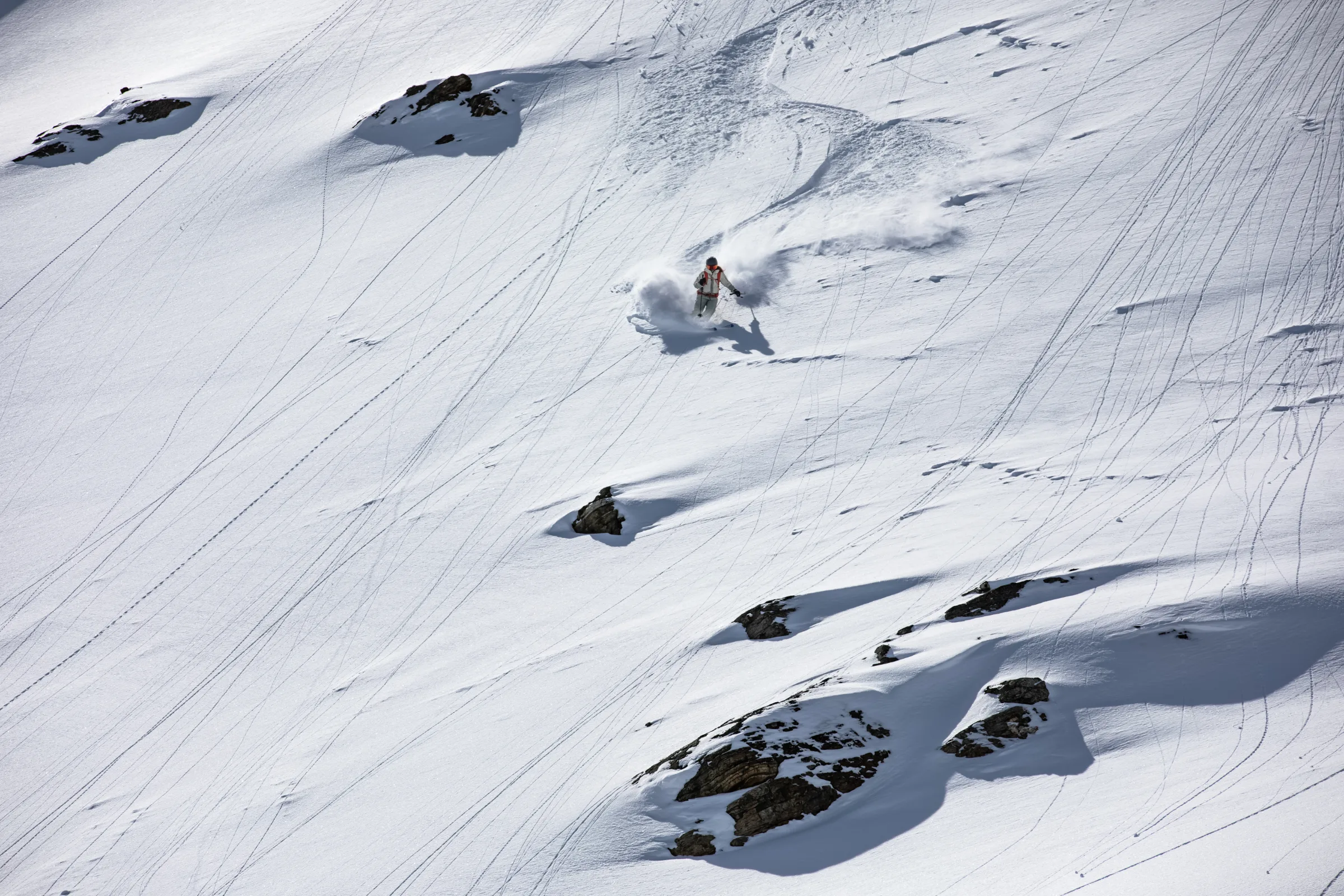 Eine Skifahrerin zieht auf einem weiten, von feinen Spuren durchzogenen Schneehang zwischen Felsen einen Schwung und hinterlässt eine breite Schneefahne.