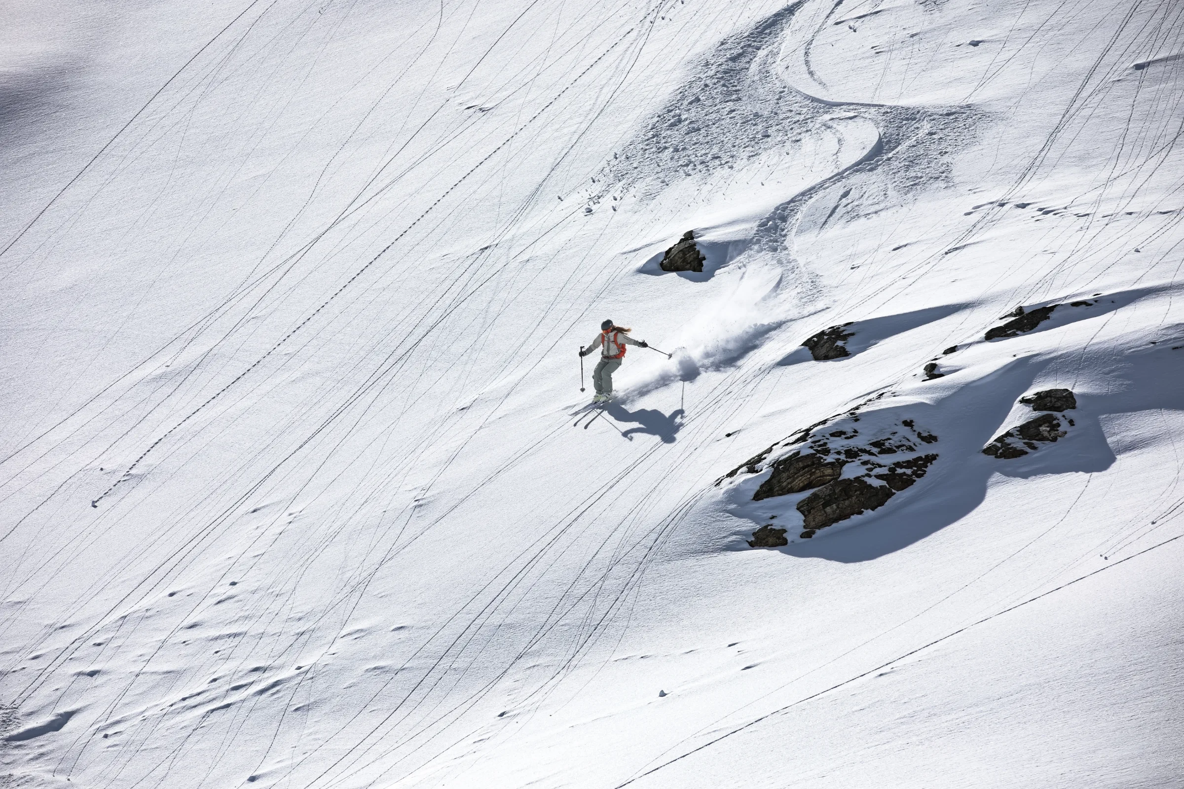 Eine Skifahrerin fährt auf einem weitläufigen Schneehang links an verschneiten Felsen vorbei und hinterlässt eine Schneefahne unterhalb ihrer geschwungenen Spur.
