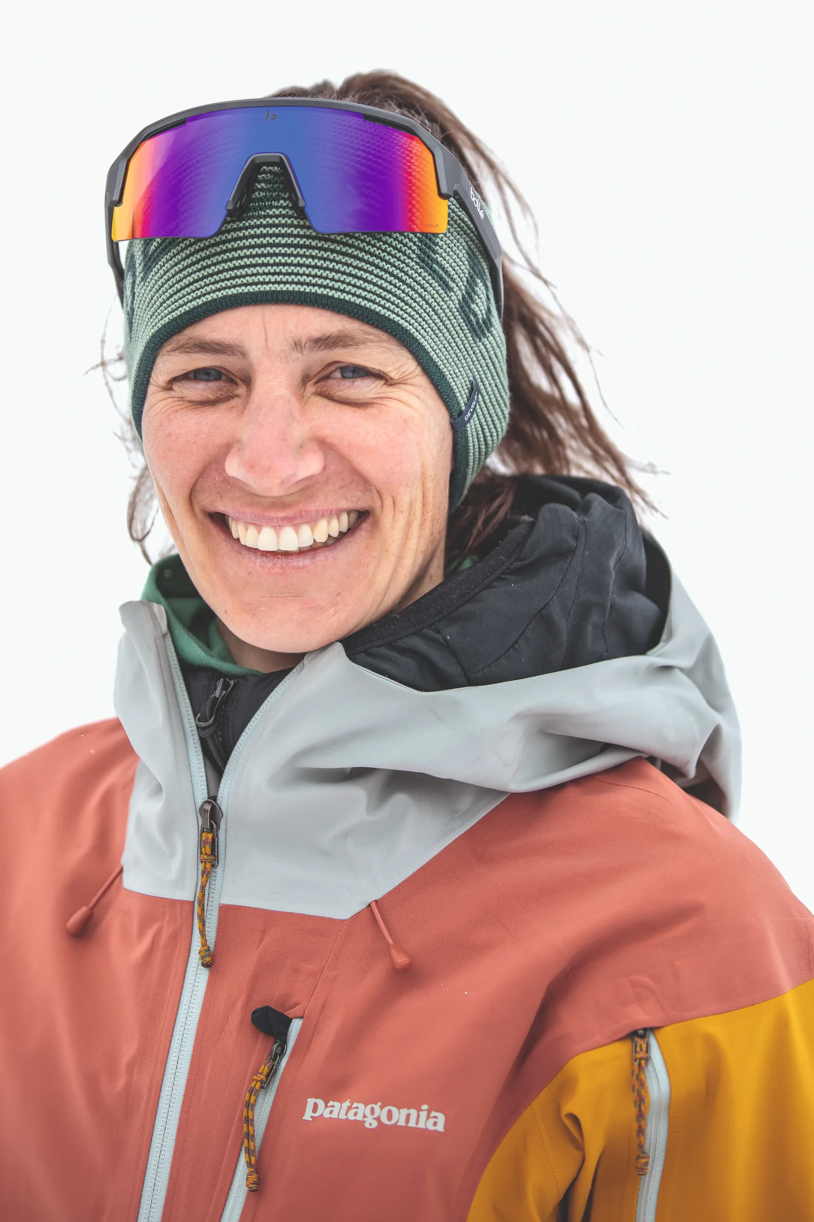 Sabine Speidel lächelt in Nahaufnahme vor weißem Hintergrund, mit grünem Strickstirnband und verspiegelter Sportbrille auf der Stirn.