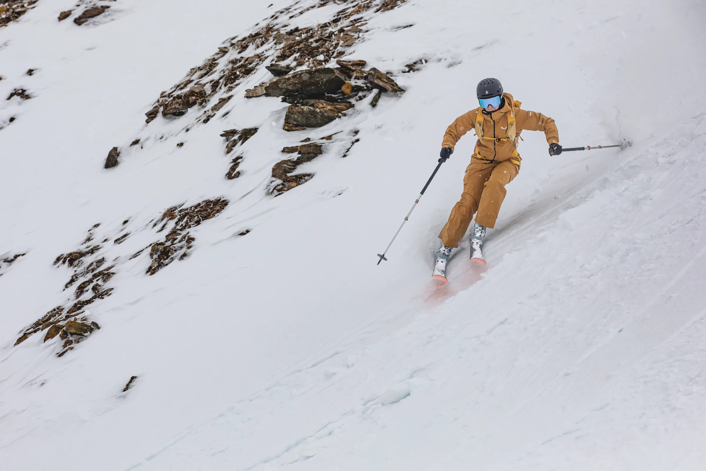 Sabine Speidel fährt in ockerfarbener Kleidung mit weit gehaltenen Skistöcken einen felsdurchsetzten Schneehang in Lenzerheide hinab.