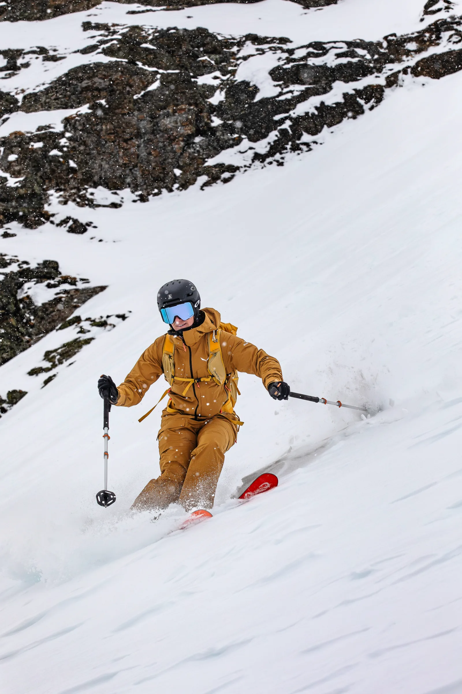 Sabine Speidel fährt mit schwarzem Helm und blauer Skibrille durch aufspritzenden Schnee einen steilen Hang unterhalb schneebedeckter Felsen hinab.