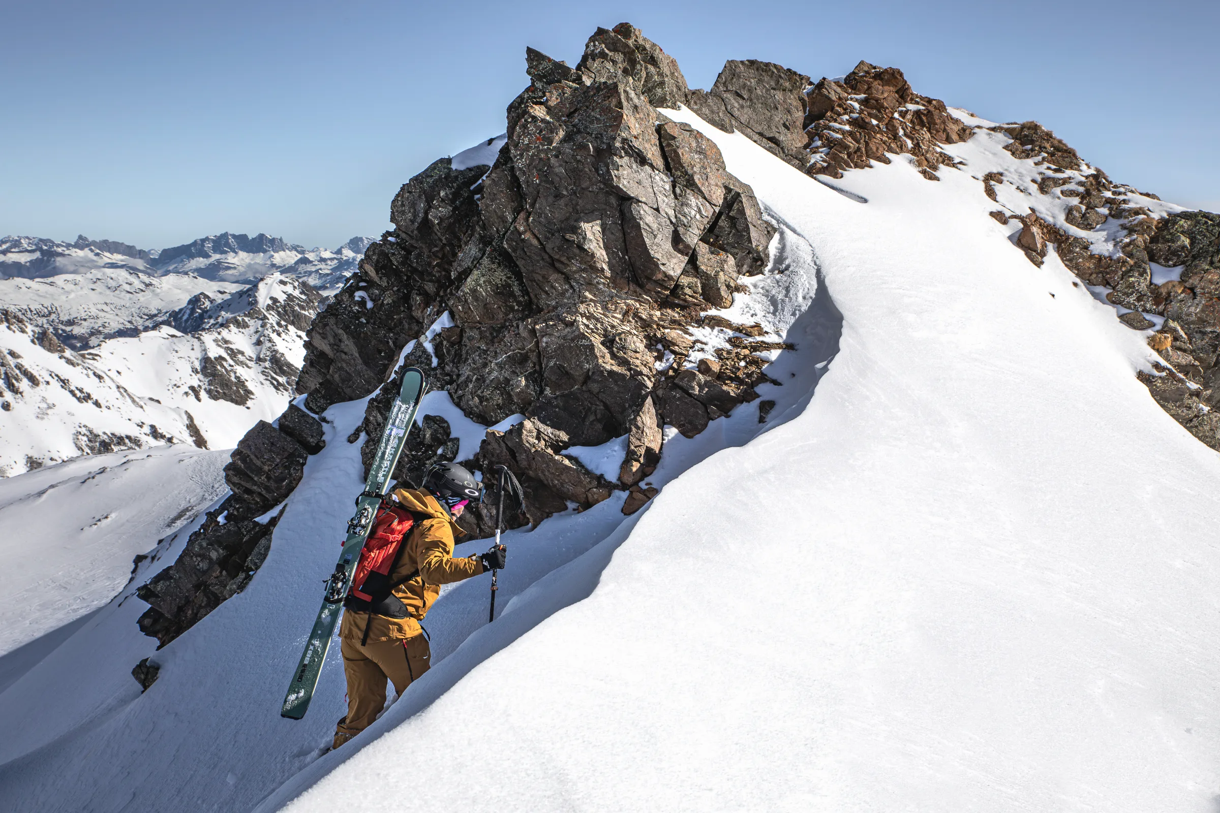 Sabine Speidel steigt mit Ski am roten Rucksack durch eine schmale Schneerinne neben einem Felsgrat auf, dahinter erstrecken sich verschneite Berge.
