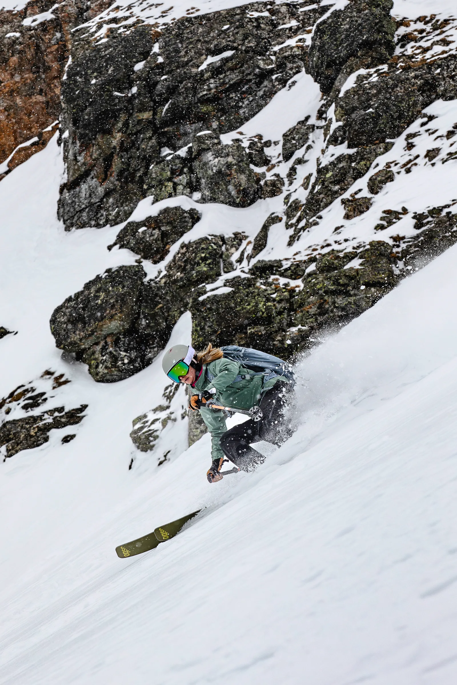 Sabrina Stickl fährt tief in der Hocke durch den Schnee vor einer Felswand, eine Hand knapp über dem Hang und eine Schneefahne hinter sich.