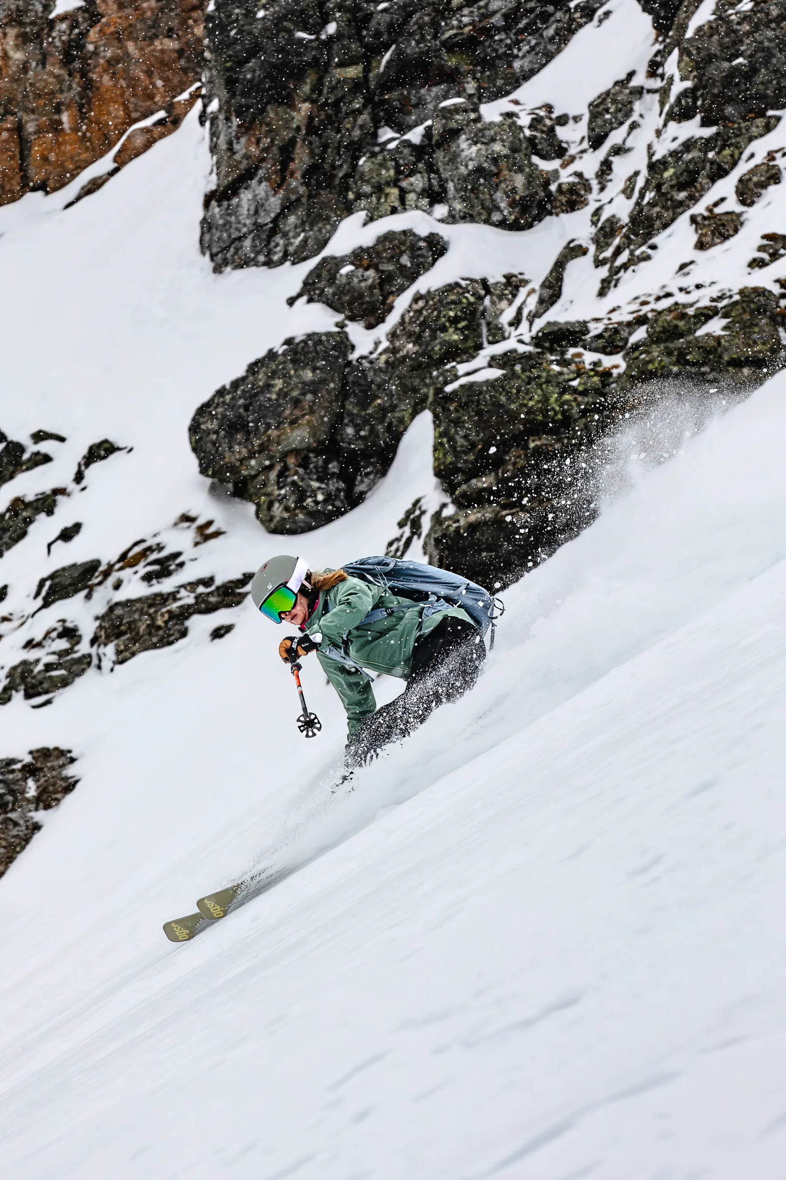 Sabrina Stickl zieht vor schneebedeckten Felsen einen tiefen Schwung, wobei aufspritzender Schnee ihre Beine teilweise verdeckt.