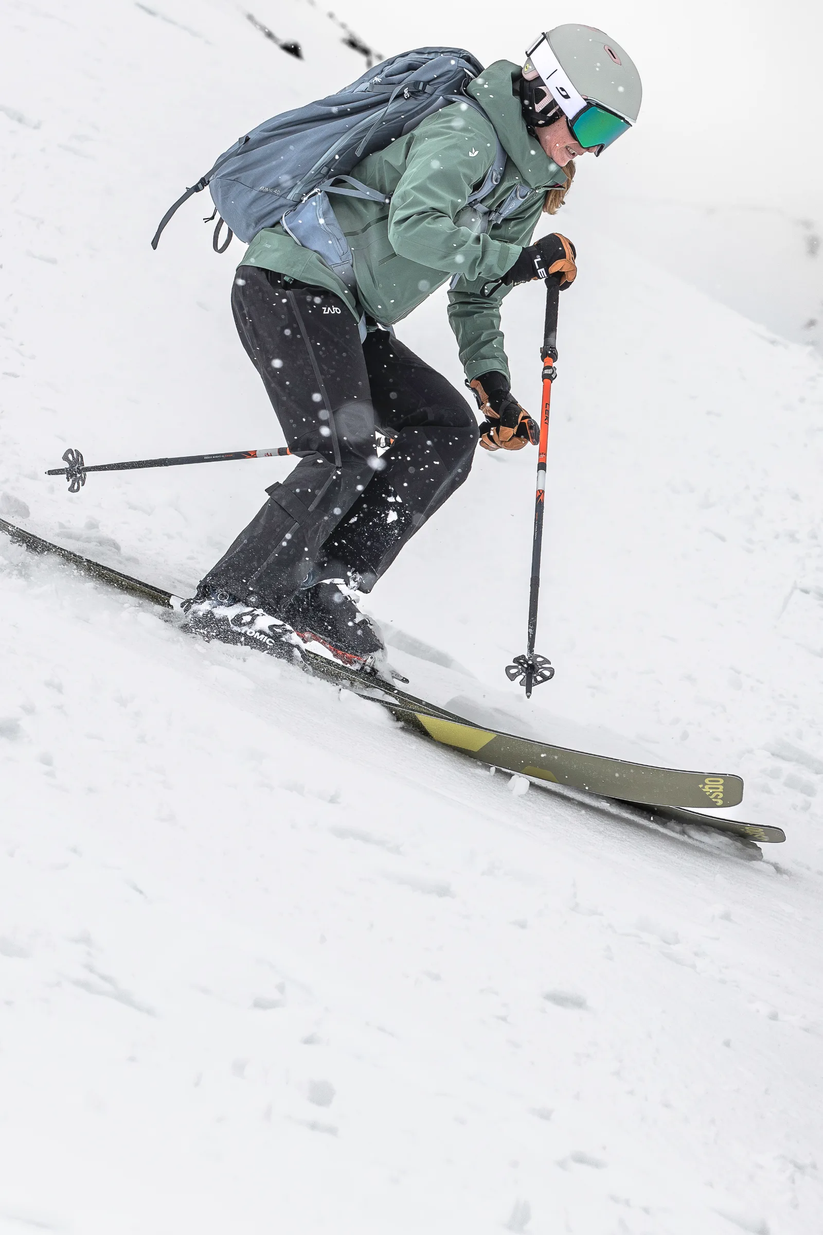 In Nahansicht fährt Sabrina Stickl mit parallelen Ski und gebeugten Knien über den Schneehang; sie trägt einen grauen Rucksack und eine verspiegelte Skibrille.