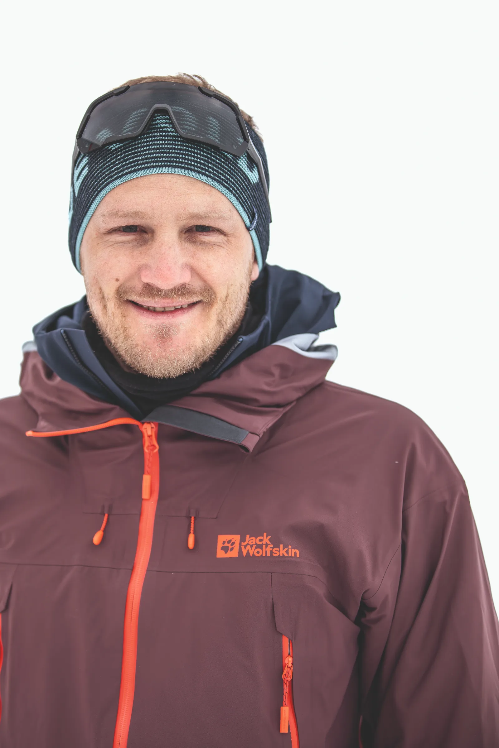 Simon Stickl lächelt vor weißem Hintergrund in die Kamera, mit gestreiftem Stirnband, hochgeschobener Sonnenbrille und burgunderfarbener Jacke.