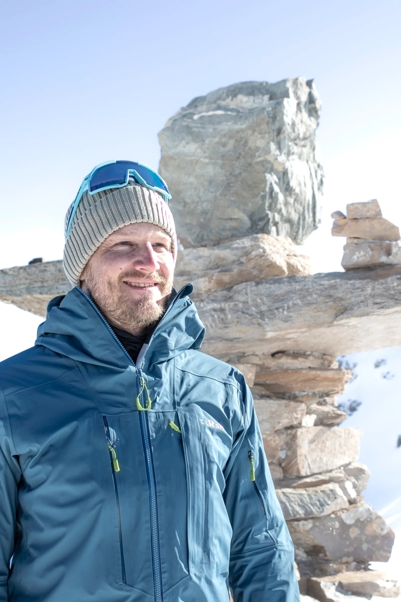 Simon Stickl lächelt in blauer Jacke und grauer Strickmütze mit aufgesetzter Sonnenbrille vor aufgeschichteten Steinplatten im Schnee.