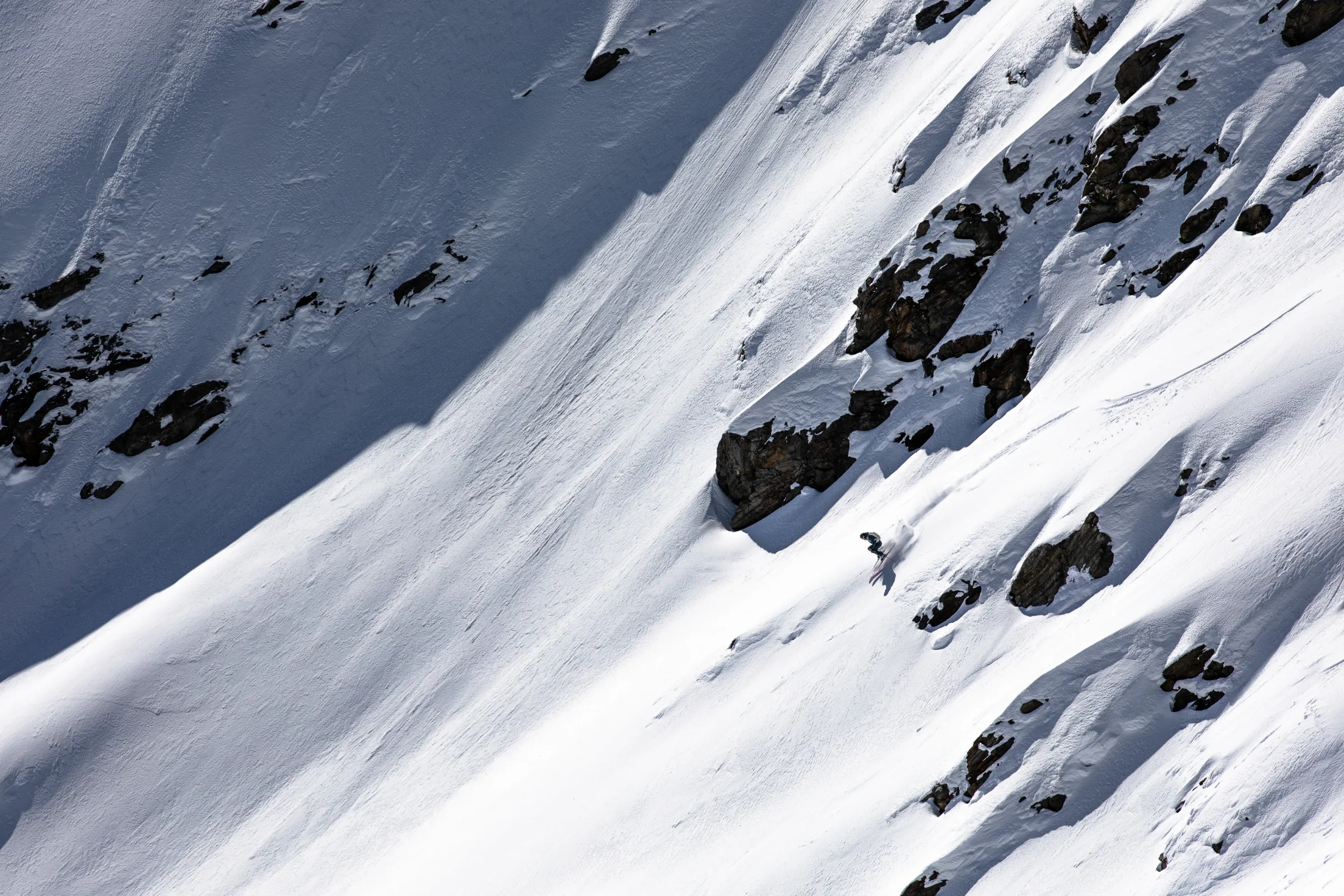 Simon Stickl fährt mit gebeugten Knien zwischen Felsen einen steilen Schneehang hinab, während hinter seinen Ski Schnee aufstäubt.