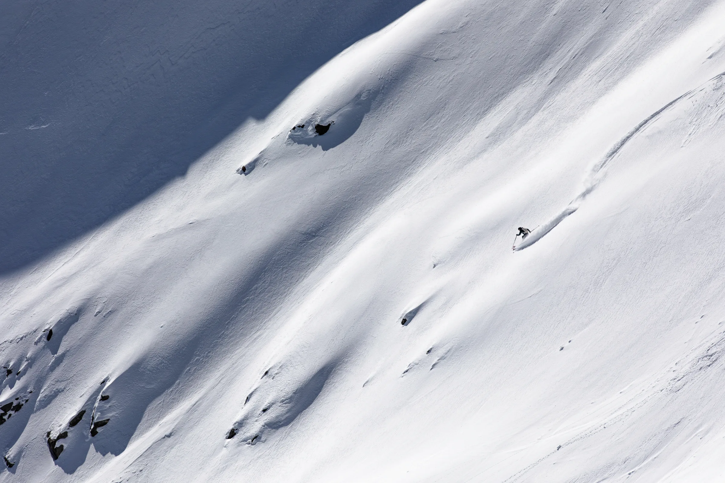 Simon Stickl fährt über einen weiten Schneehang mit einzelnen Felsen; seine lange Spur zieht sich nach rechts oben durch den Schnee.