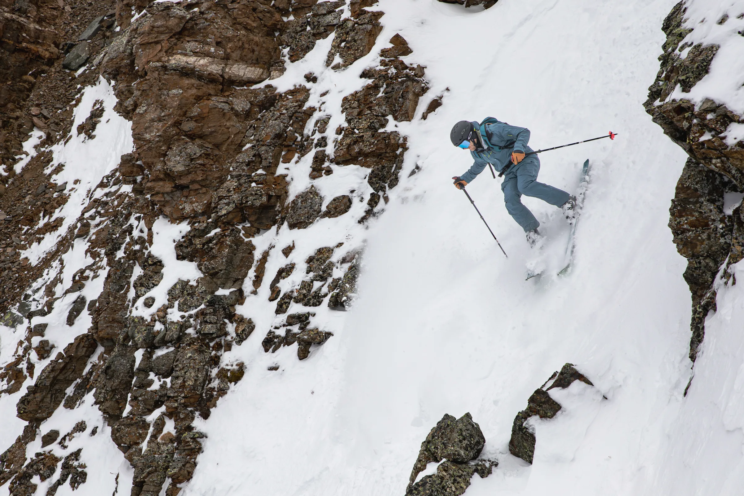 Simon Stickl fährt in blauer Skikleidung mit Helm, Skibrille und Rucksack durch eine enge Schneerinne zwischen braunen Felswänden.