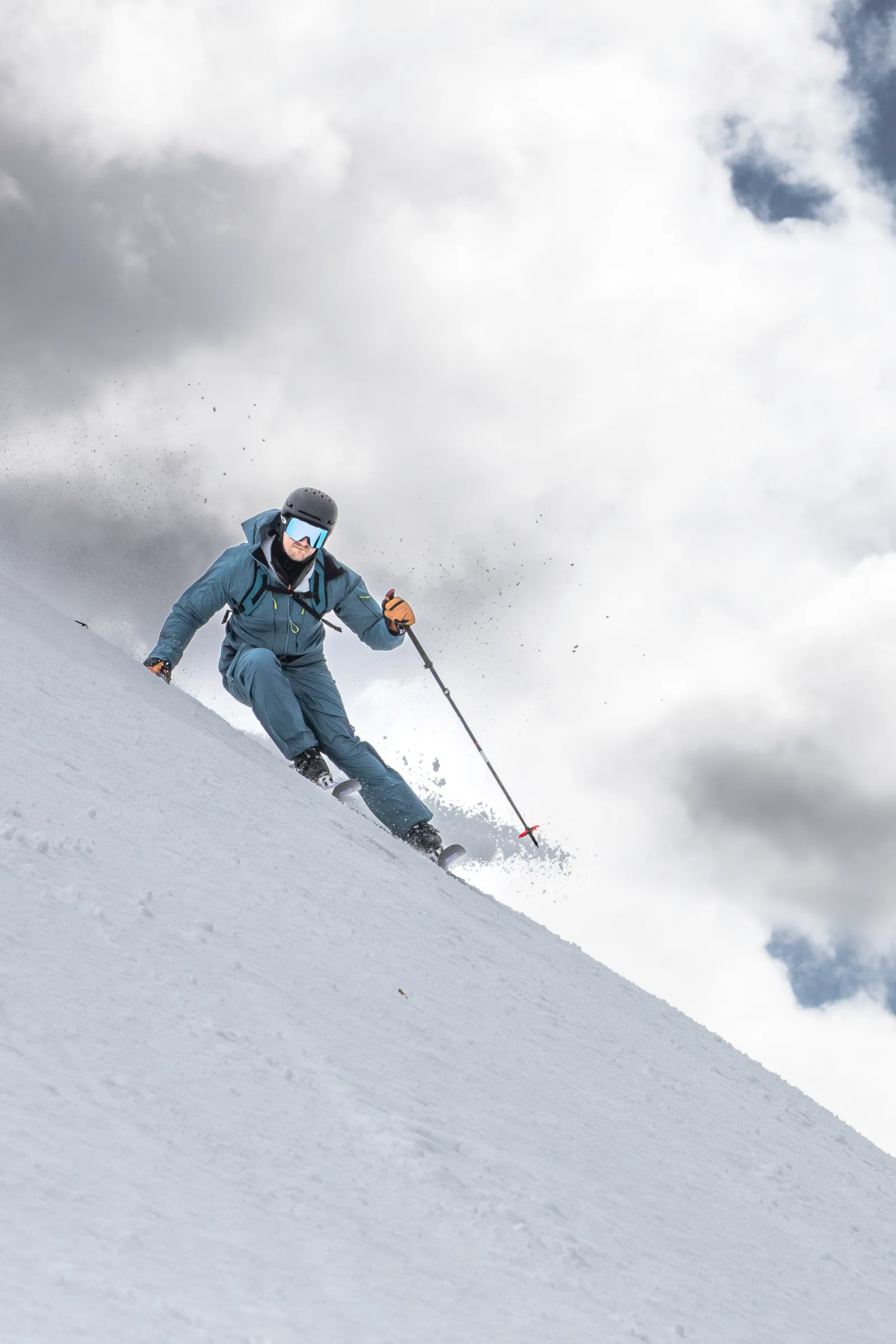 Aus tiefer Perspektive fährt Simon Stickl mit gebeugten Knien einen Schneehang hinab; Schnee spritzt vor dem wolkigen Himmel auf.