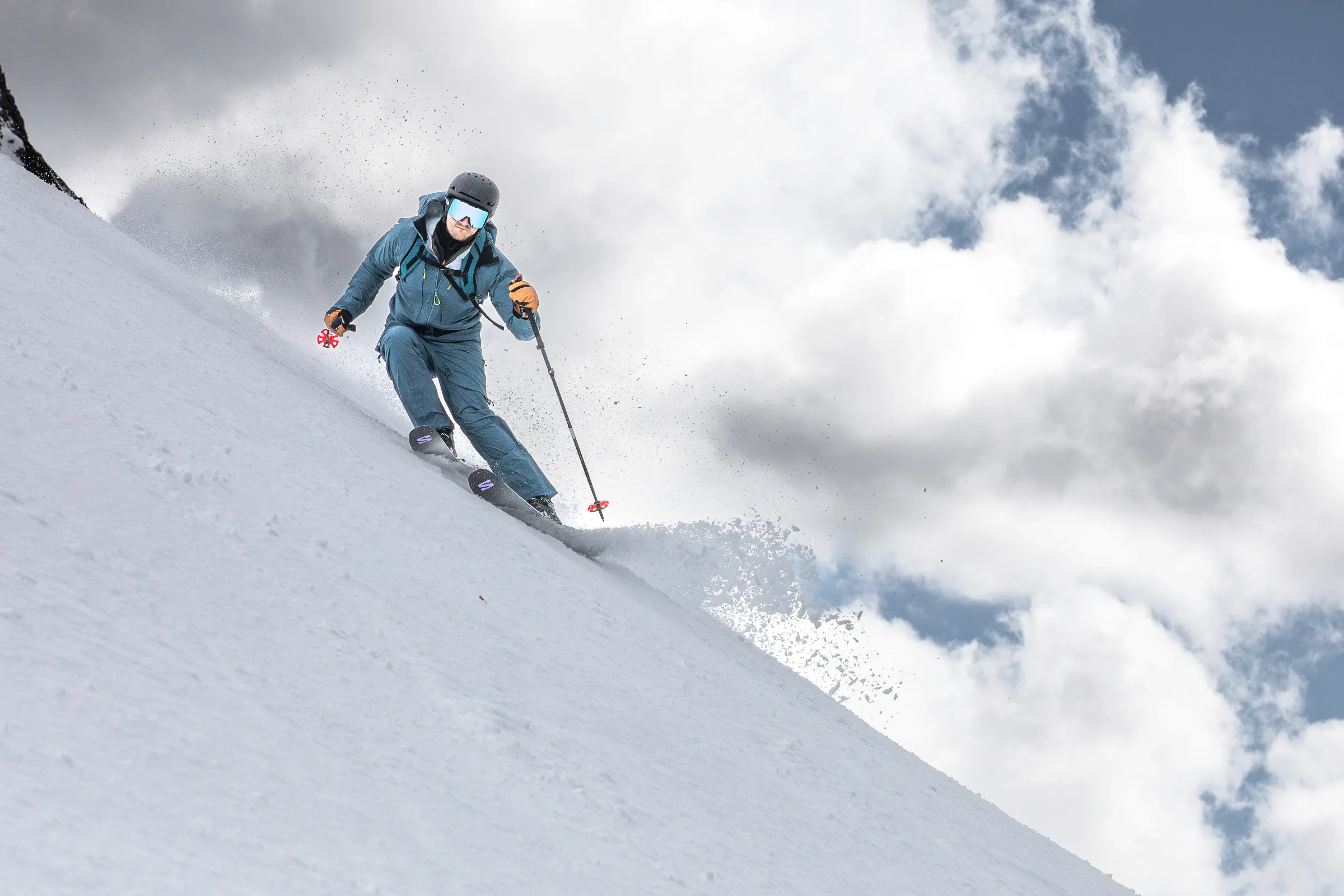Simon Stickl kantet seine Ski am steilen Schneehang auf und hält die Stöcke seitlich, während vor dem wolkigen Himmel Schnee aufwirbelt.