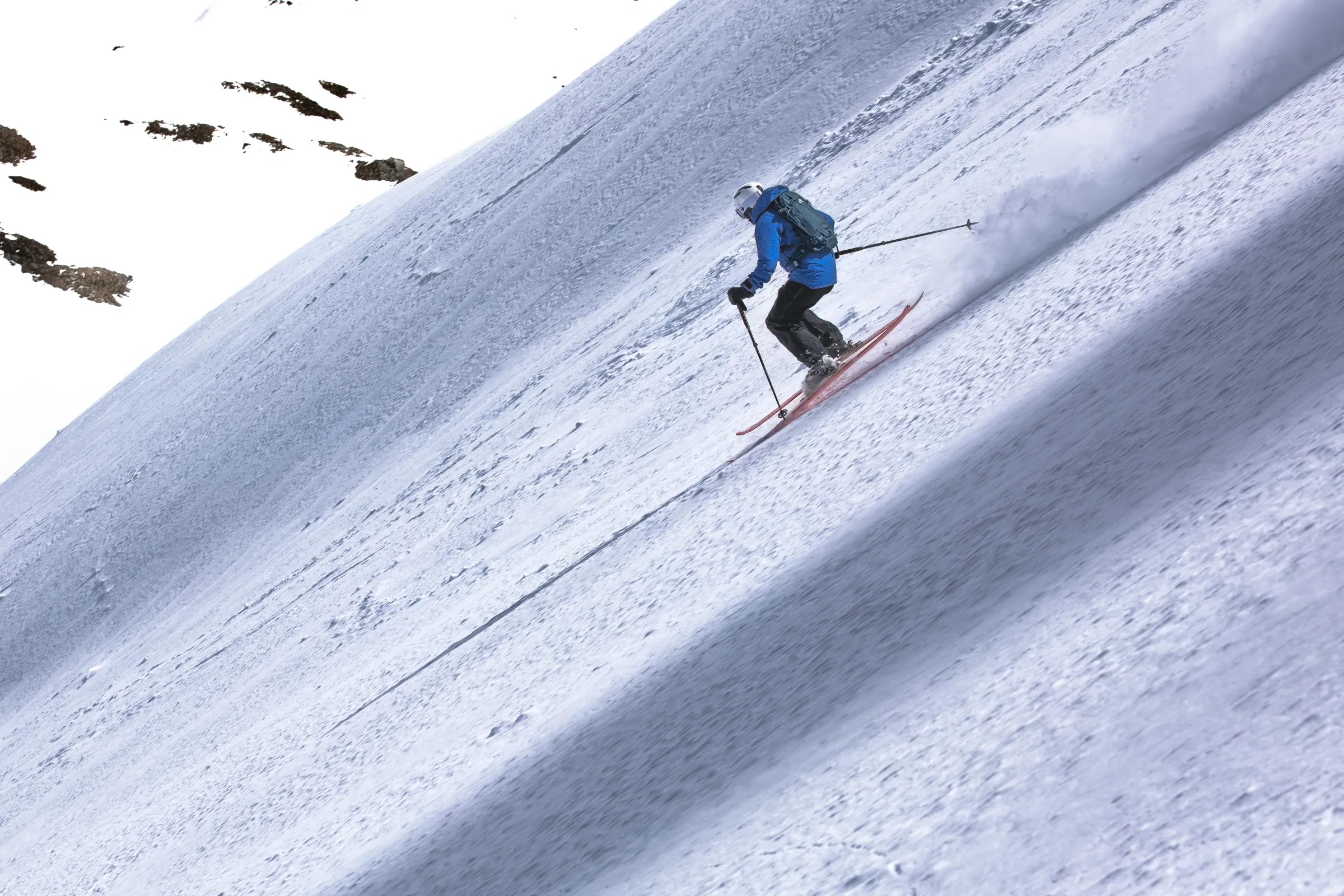 Von hinten gesehen fährt Simon Stickl mit Rucksack auf roten Ski einen steilen Hang hinab und zieht eine Schneefahne hinter sich her; links ragen Felsen aus dem Schnee.