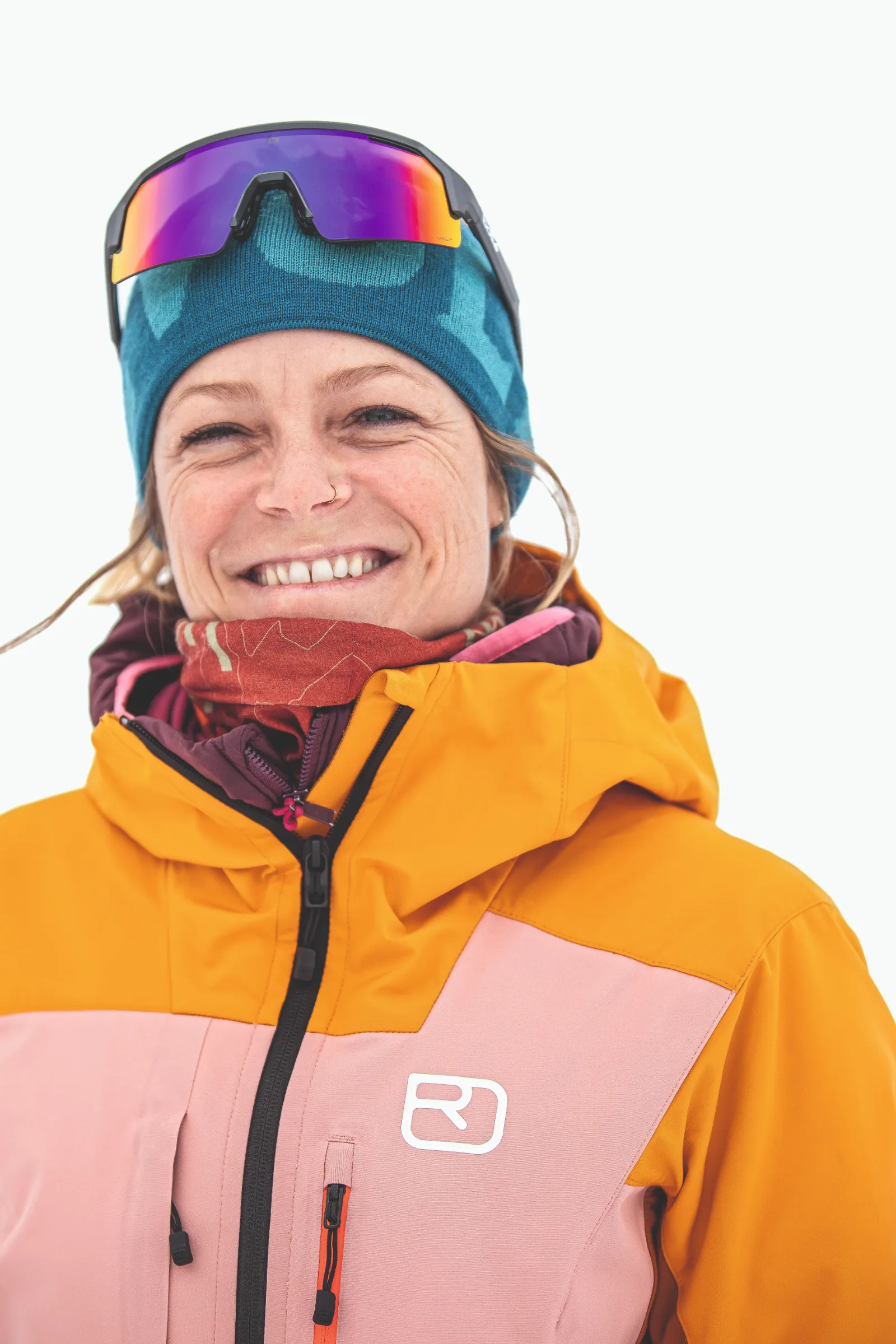 Sofia Muigg lächelt vor weißem Hintergrund mit blauer Mütze, darauf sitzender verspiegelter Sportbrille und orange-rosa Skijacke.