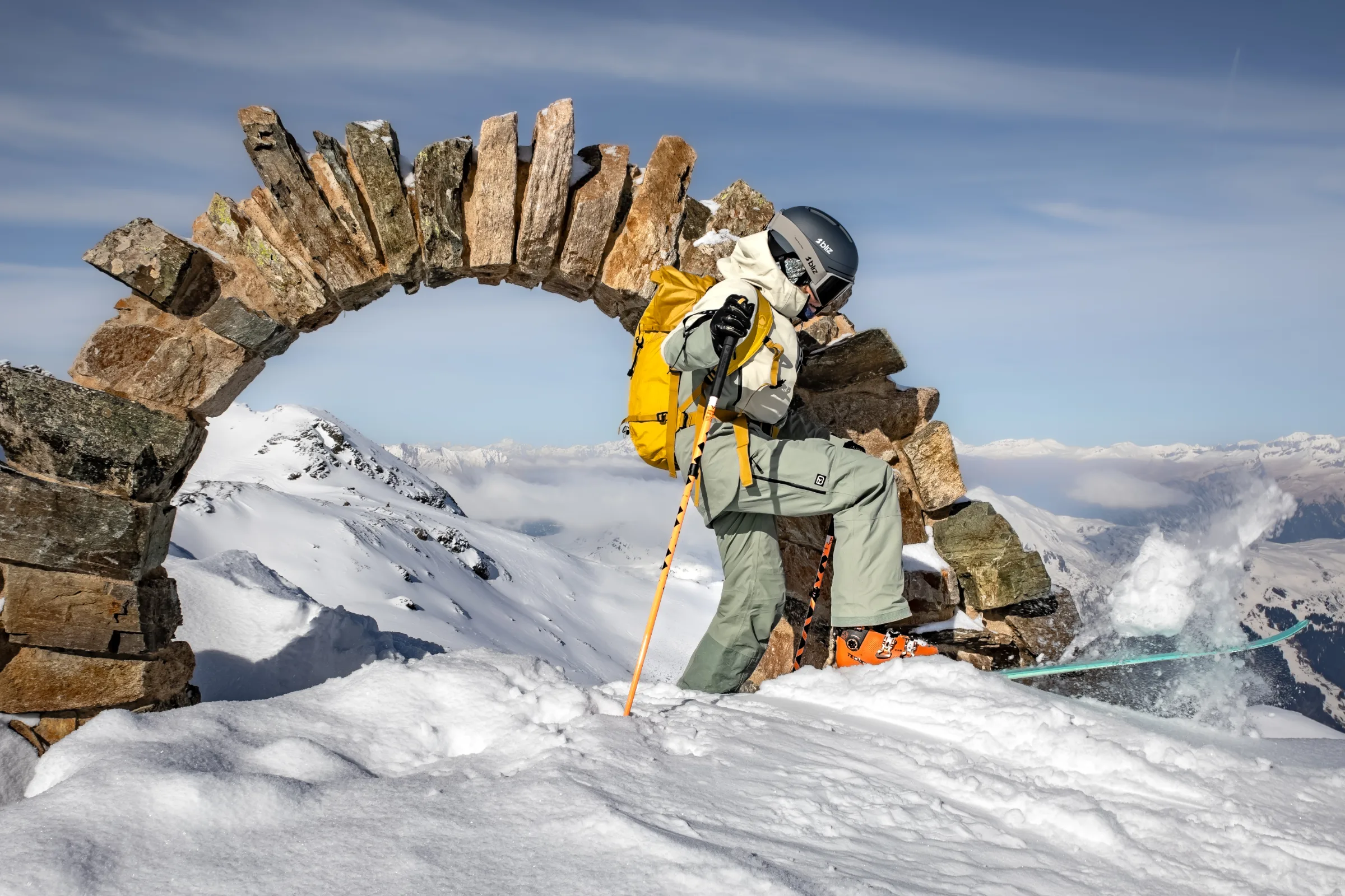 Sofia Muigg hebt neben einem Steinbogen einen Ski an und wirbelt Schnee auf, mit gelbem Rucksack und verschneiten Bergen von Lenzerheide im Hintergrund.