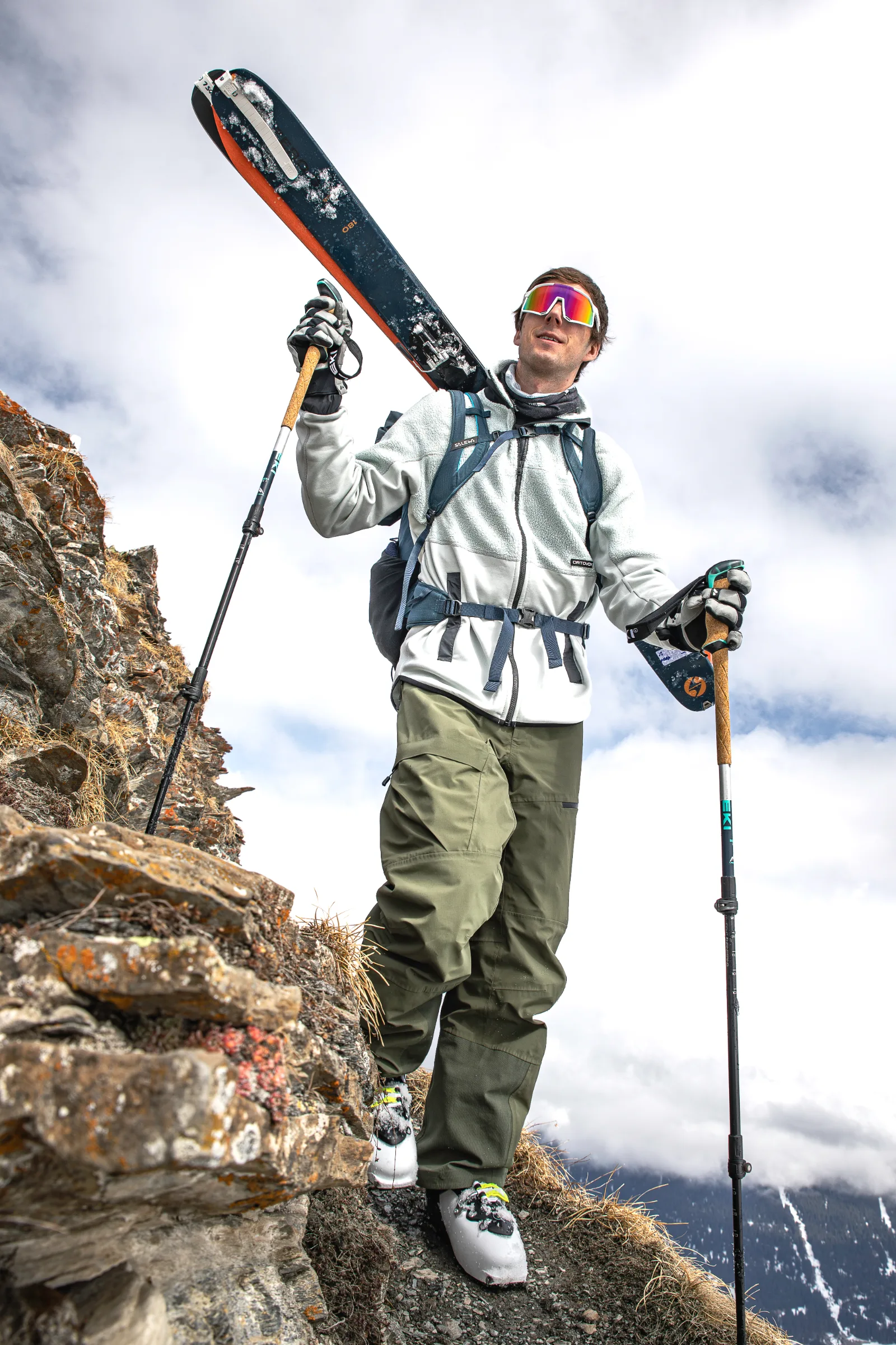 Valentin Rapp geht mit Ski am Rucksack und Stöcken in den Händen über einen schmalen felsigen Bergpfad unter bewölktem Himmel.